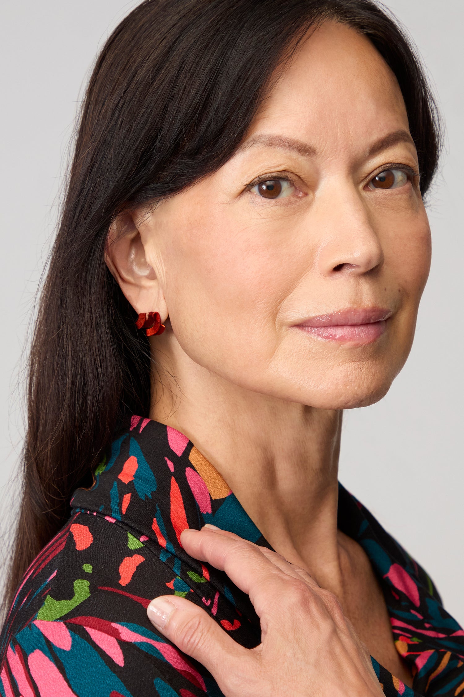 A woman with straight dark hair wears a patterned shirt and Ribbon Earrings made of red anodised aluminium, looking at the camera with a neutral expression and her hand resting on her shoulder.