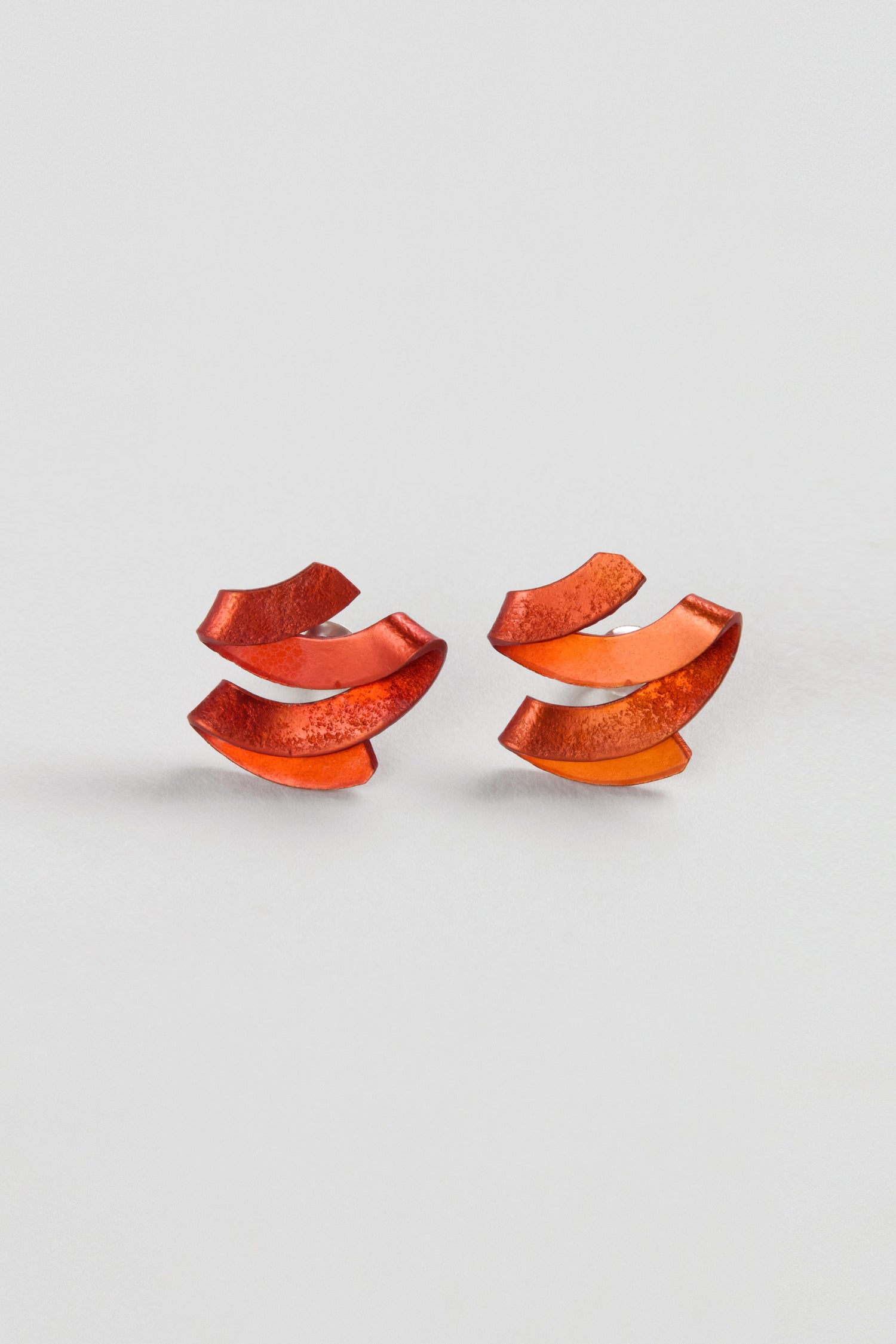 A pair of Ribbon Earrings in metallic, curved red anodised aluminium with a layered abstract design, displayed on a plain white background to showcase sculptural jewellery artistry.