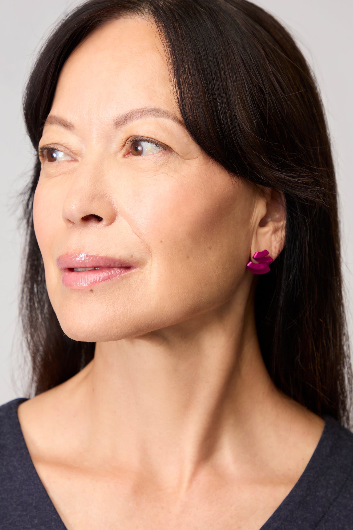 A woman with long dark hair looks to the side, wearing a dark top and pink Ribbon Earrings featuring a sculptural, flower-shaped anodised aluminium design.