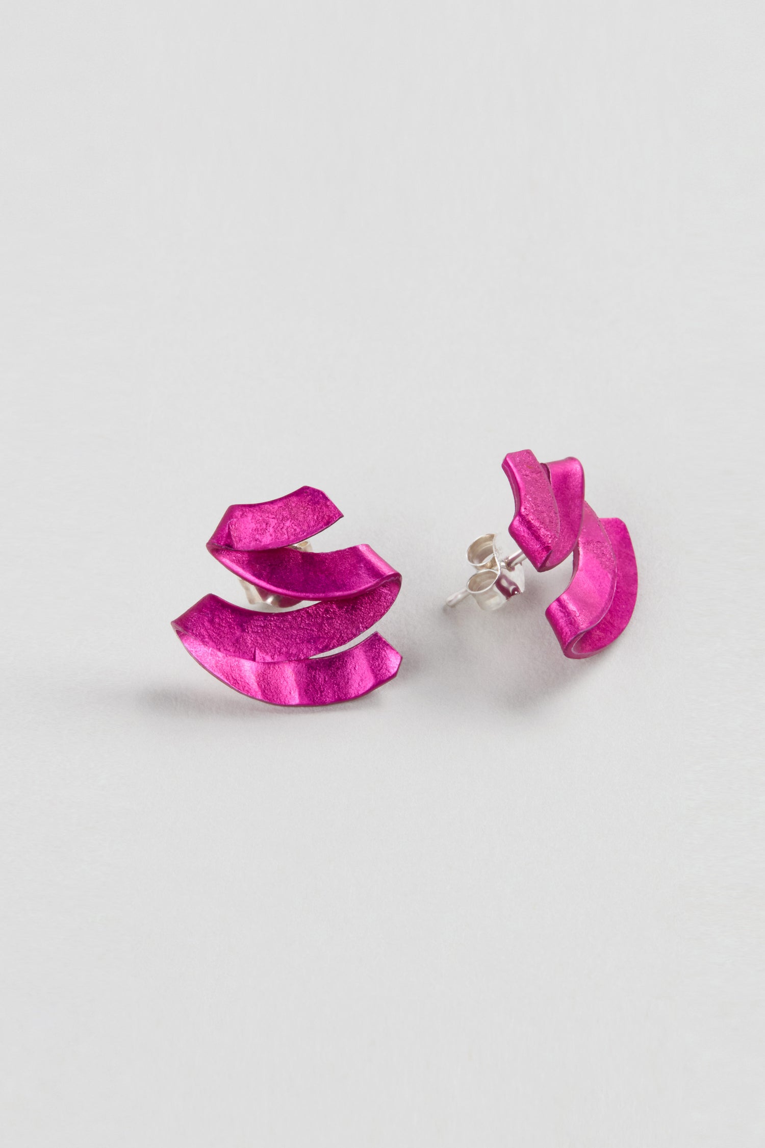 A pair of Ribbon Earrings by Oana Millet, crafted from anodised aluminium in metallic pink, are displayed on a plain light background—an elegant sculptural jewellery piece.