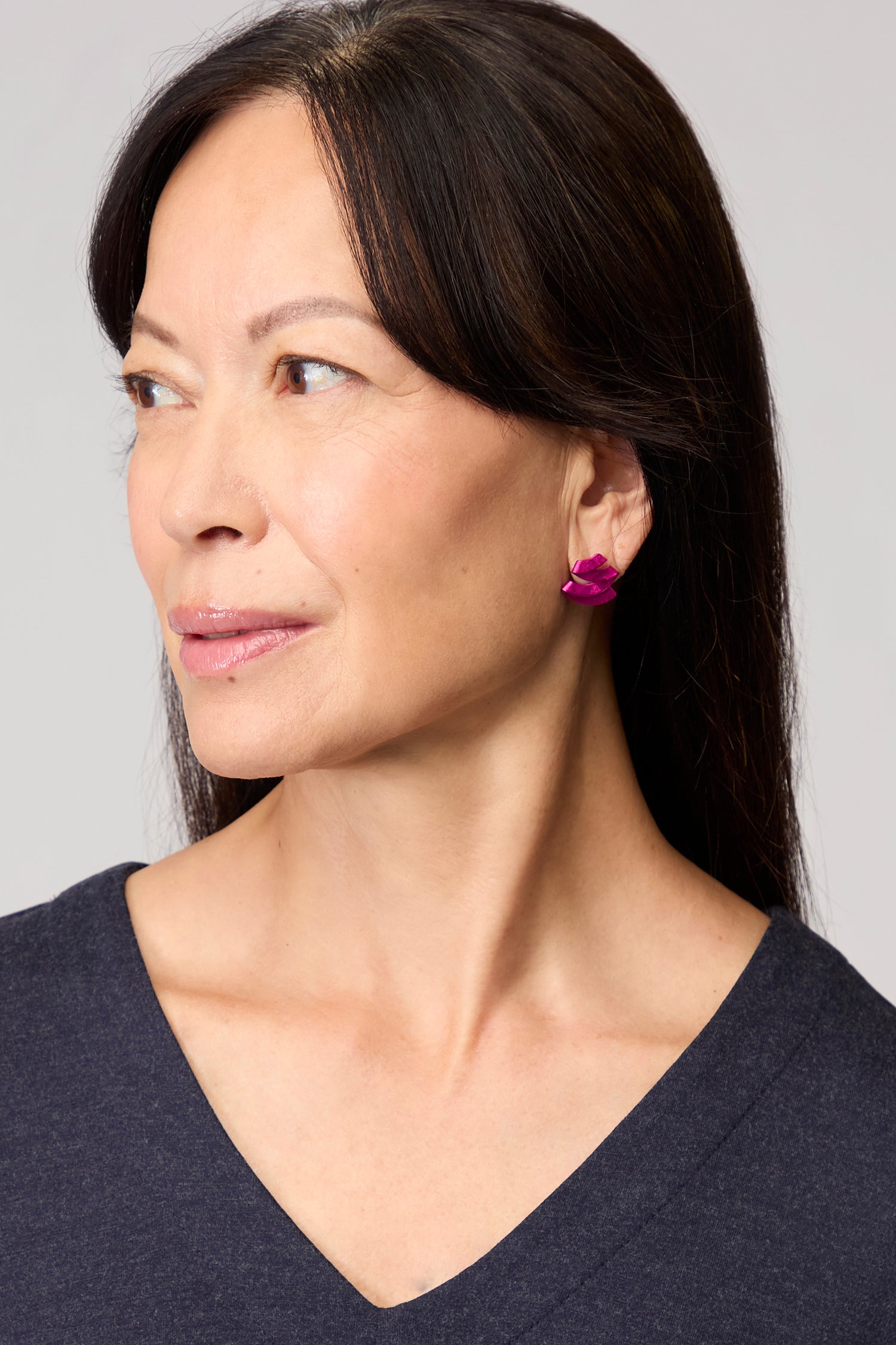 Woman with long dark hair in a dark V-neck top wears bright pink Ribbon Earrings by Oana Millet, gazing left against a light gray background—a striking touch of sculptural jewellery.