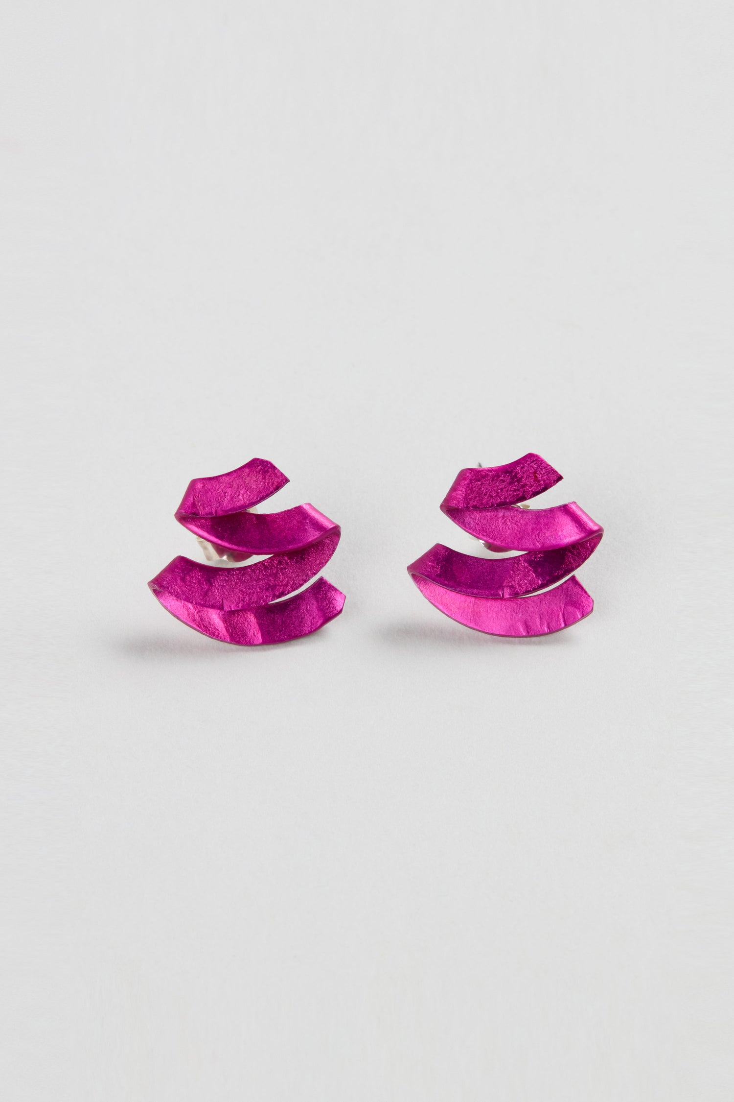 A pair of Ribbon Earrings in metallic pink, featuring a layered, abstract shape and crafted from anodised aluminium, are displayed on a plain white background to highlight their striking sculptural design.