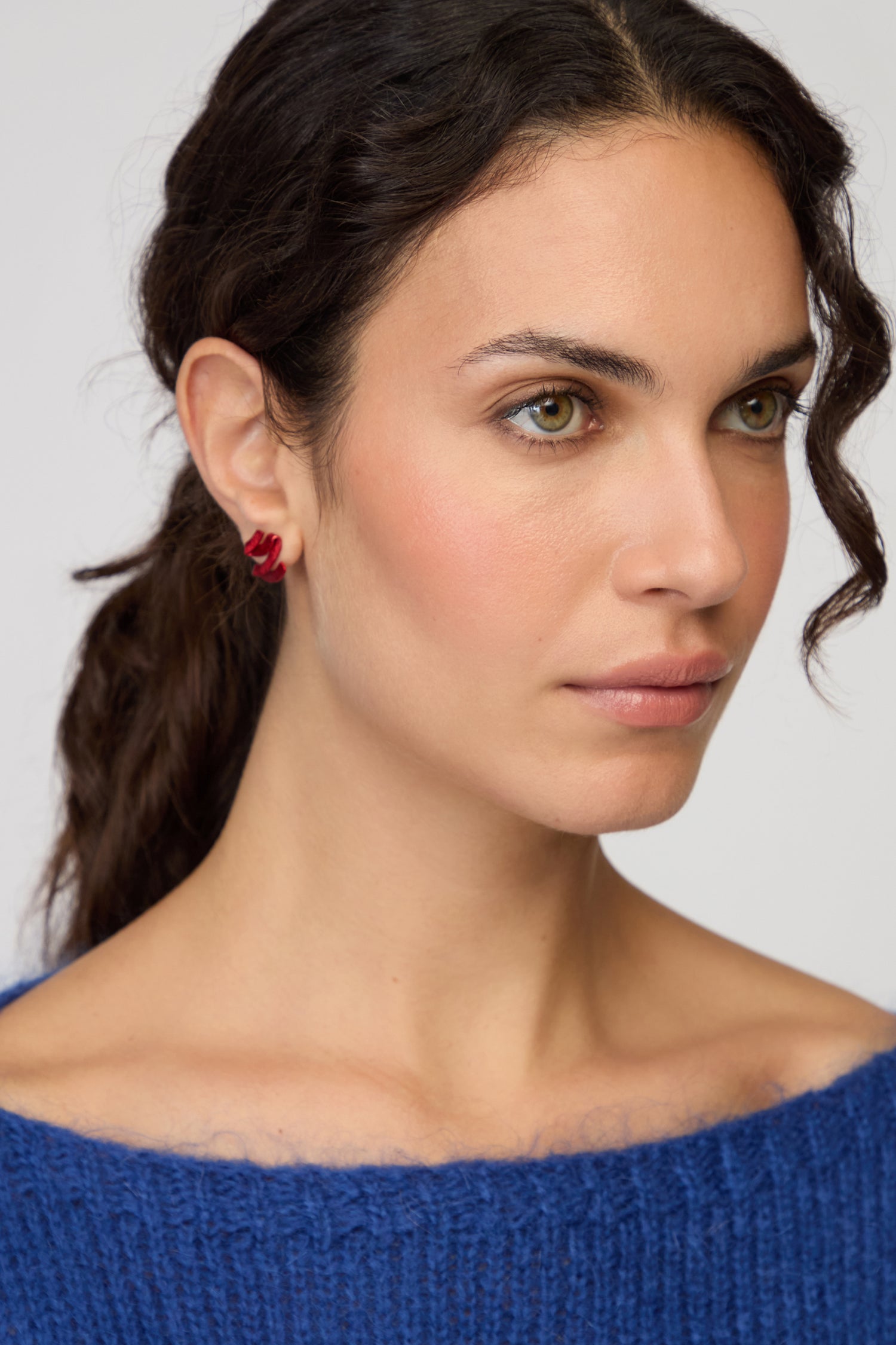 A woman with wavy dark hair in a ponytail wears a blue knit sweater and sculptural Ribbon Earrings as she gazes slightly to the side against a plain background.