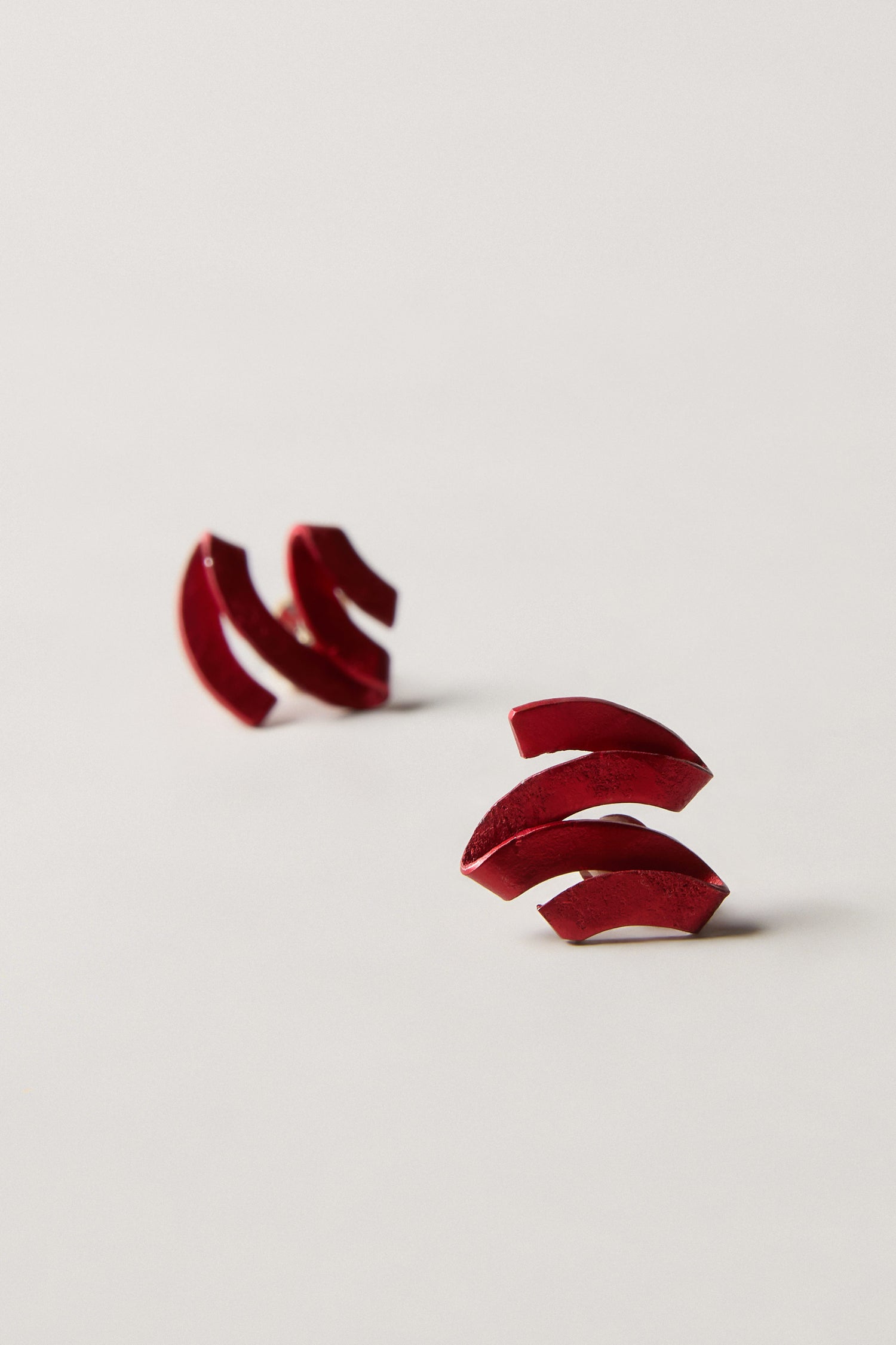 A pair of red, geometric, crescent-shaped anodised aluminium Ribbon Earrings displayed on a light background—an elegant example of sculptural jewellery.
