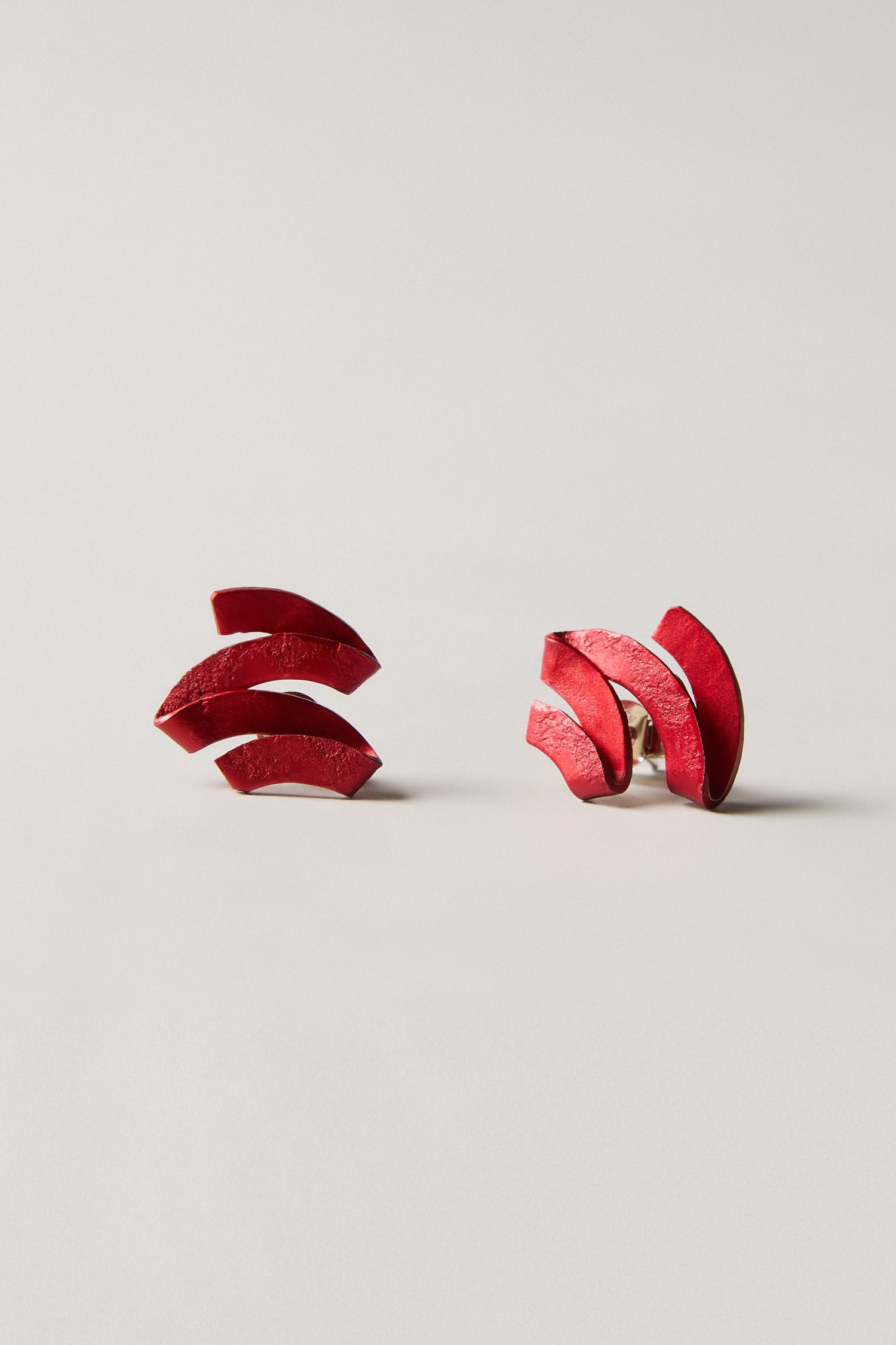 A pair of red, curved, interlocking Ribbon Earrings displayed on a plain light background—striking sculptural jewellery crafted from anodised aluminium.