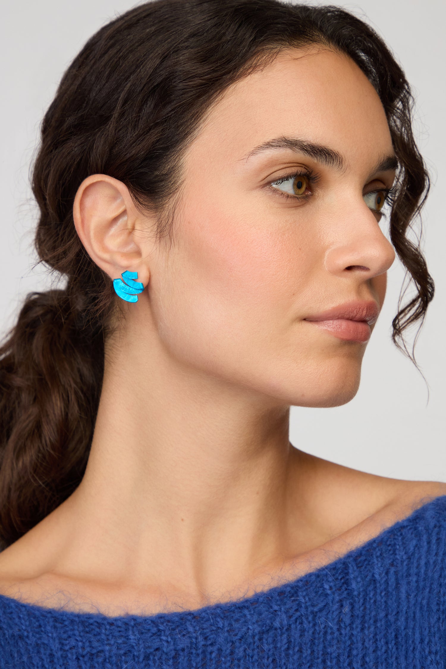 Woman with wavy brown hair, wearing a blue sweater and Ribbon Earrings made from sculptural anodised aluminium, looks to the side against a neutral background.