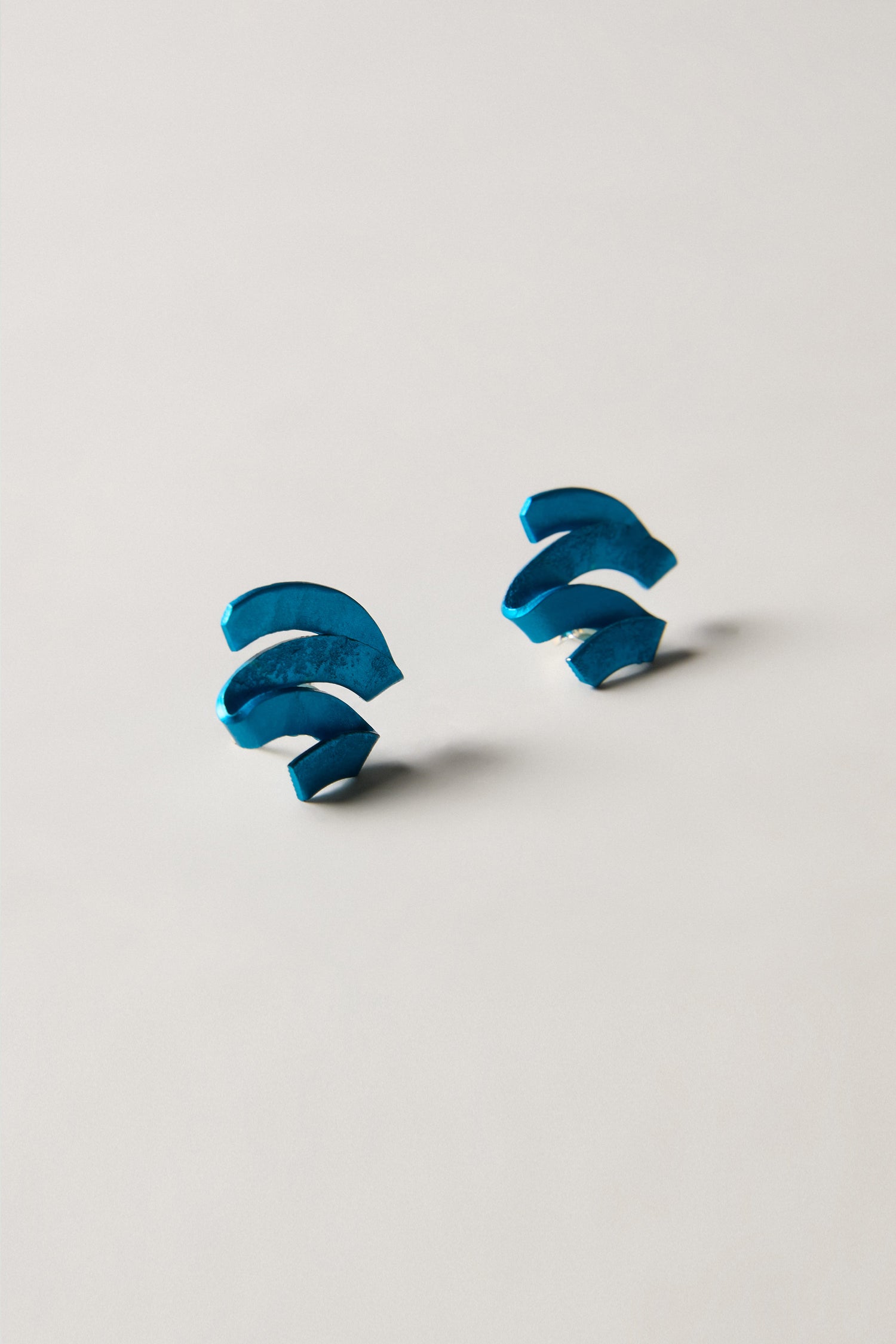 Two blue, curved, abstract-shaped objects—resembling Ribbon Earrings—are placed symmetrically on a plain light-colored surface, evoking the elegance of sculptural jewellery.
