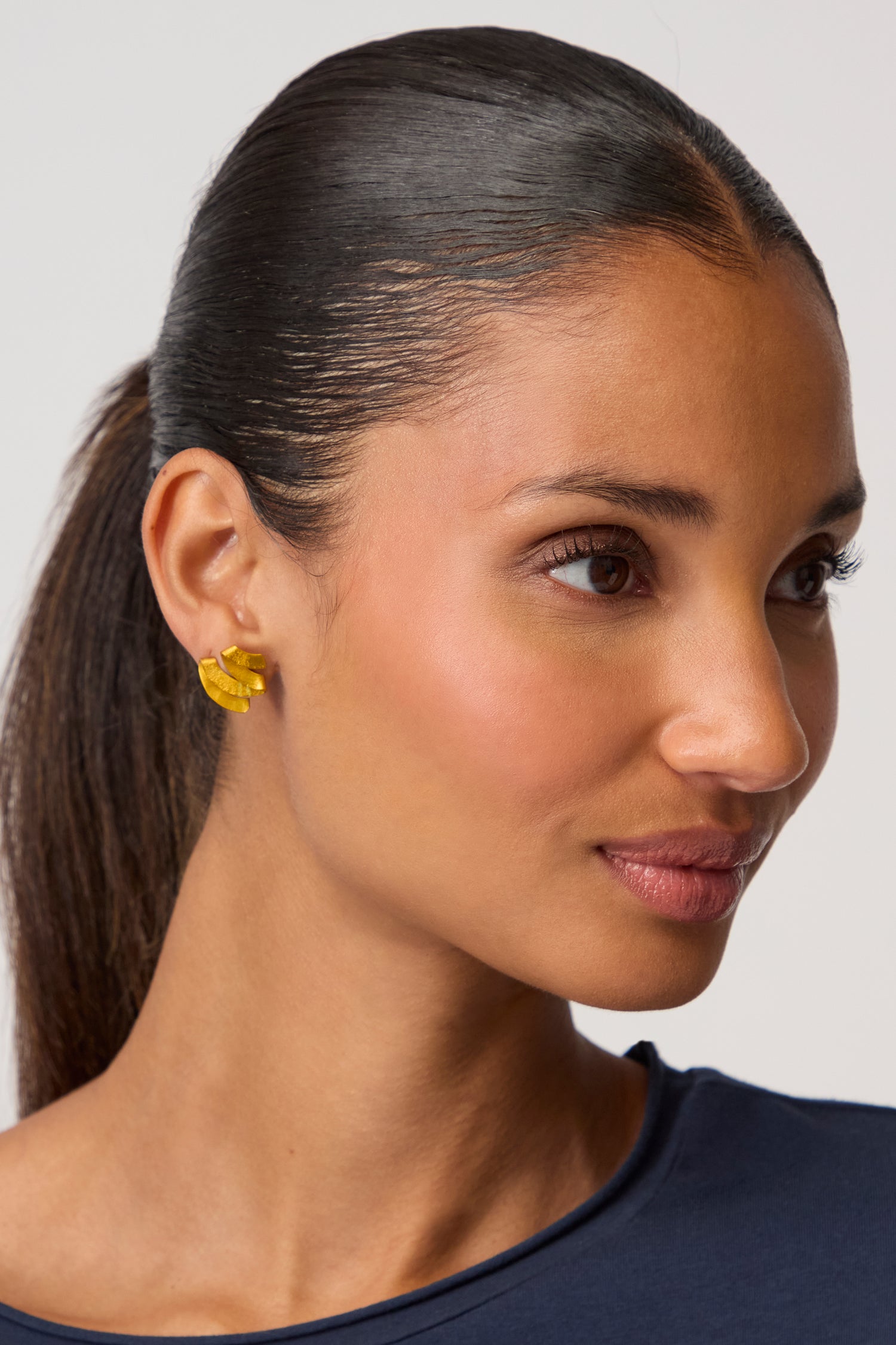 A woman with smooth, dark hair pulled back wears a navy top and striking Ribbon Earrings—sculptural jewellery crafted from anodised aluminium—as she looks to the side against a plain background.