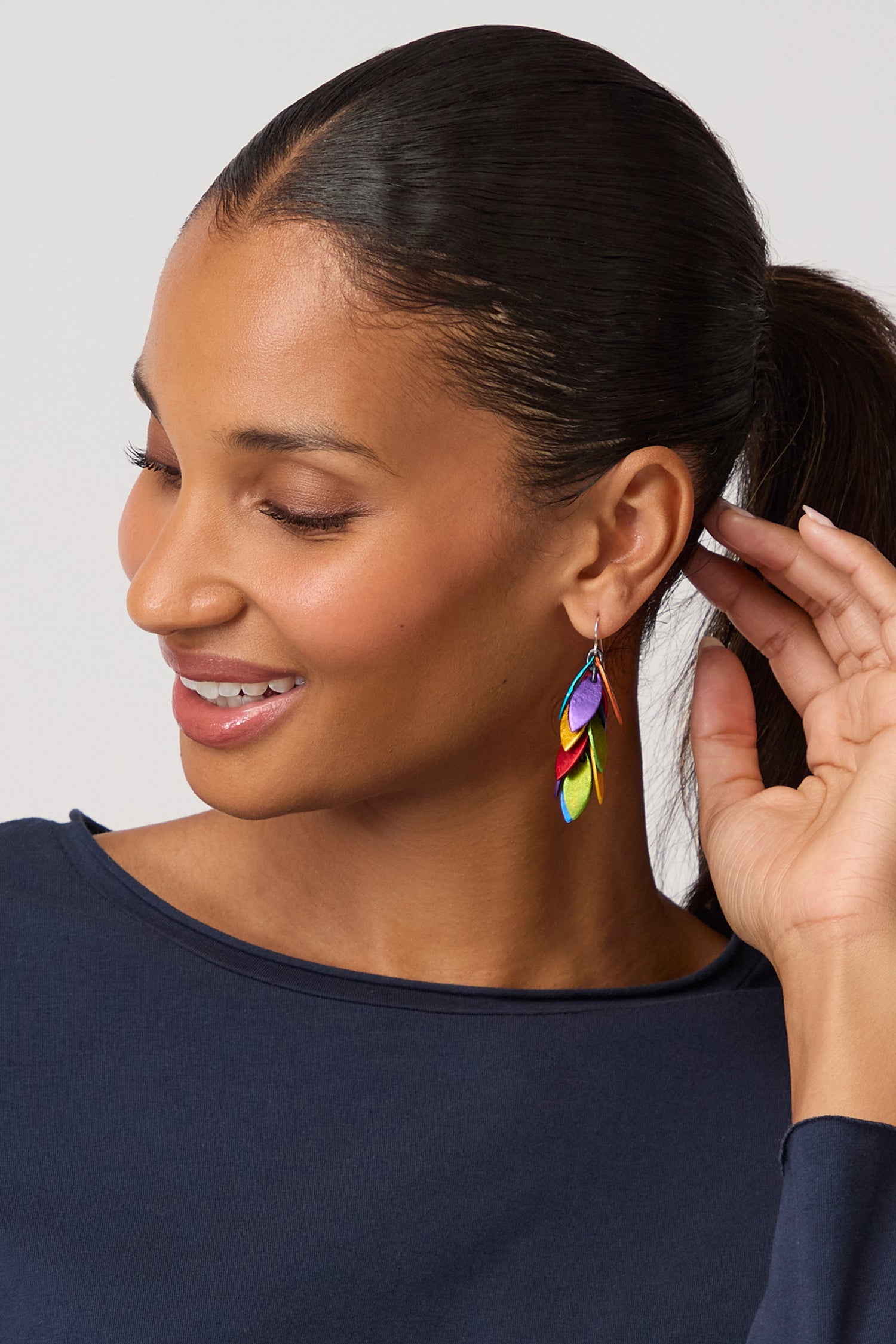 Smiling woman with a ponytail wears a navy top and Rainbow Scattered Leaves Earrings, touching her ear and looking down while showcasing her love for contemporary jewellery.