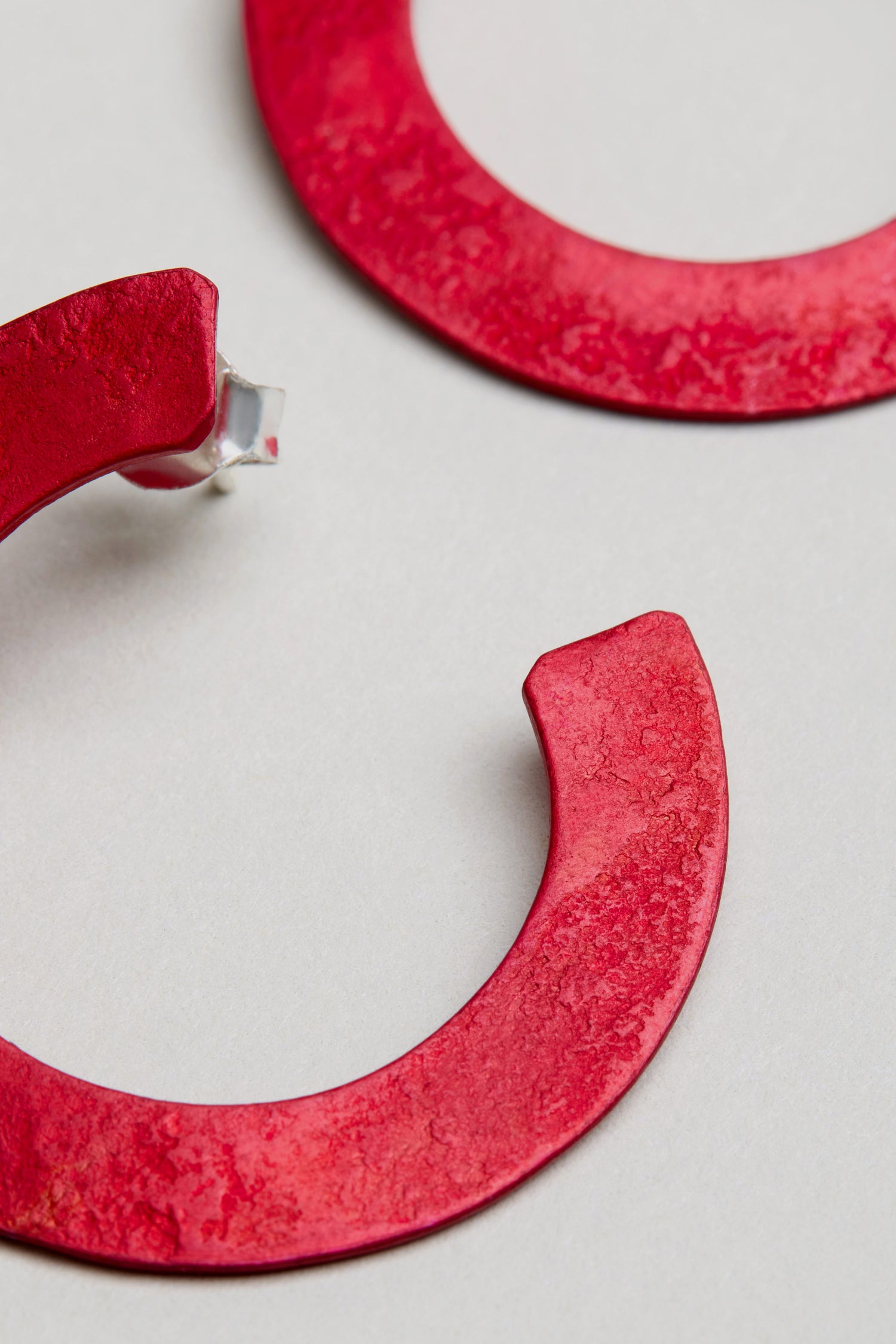 Two textured red anodised aluminium C Curve Earrings are displayed on a light surface, with one modern hoop earring visibly broken at the post.