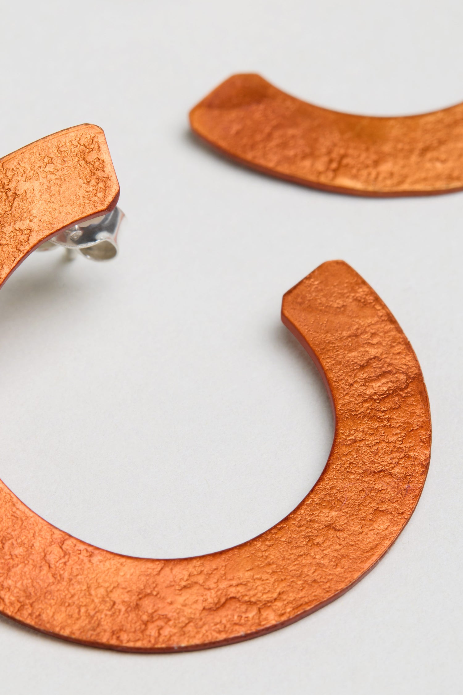 Close-up of two textured C Curve Earrings in metallic orange anodised aluminium, displayed on a light grey background.
