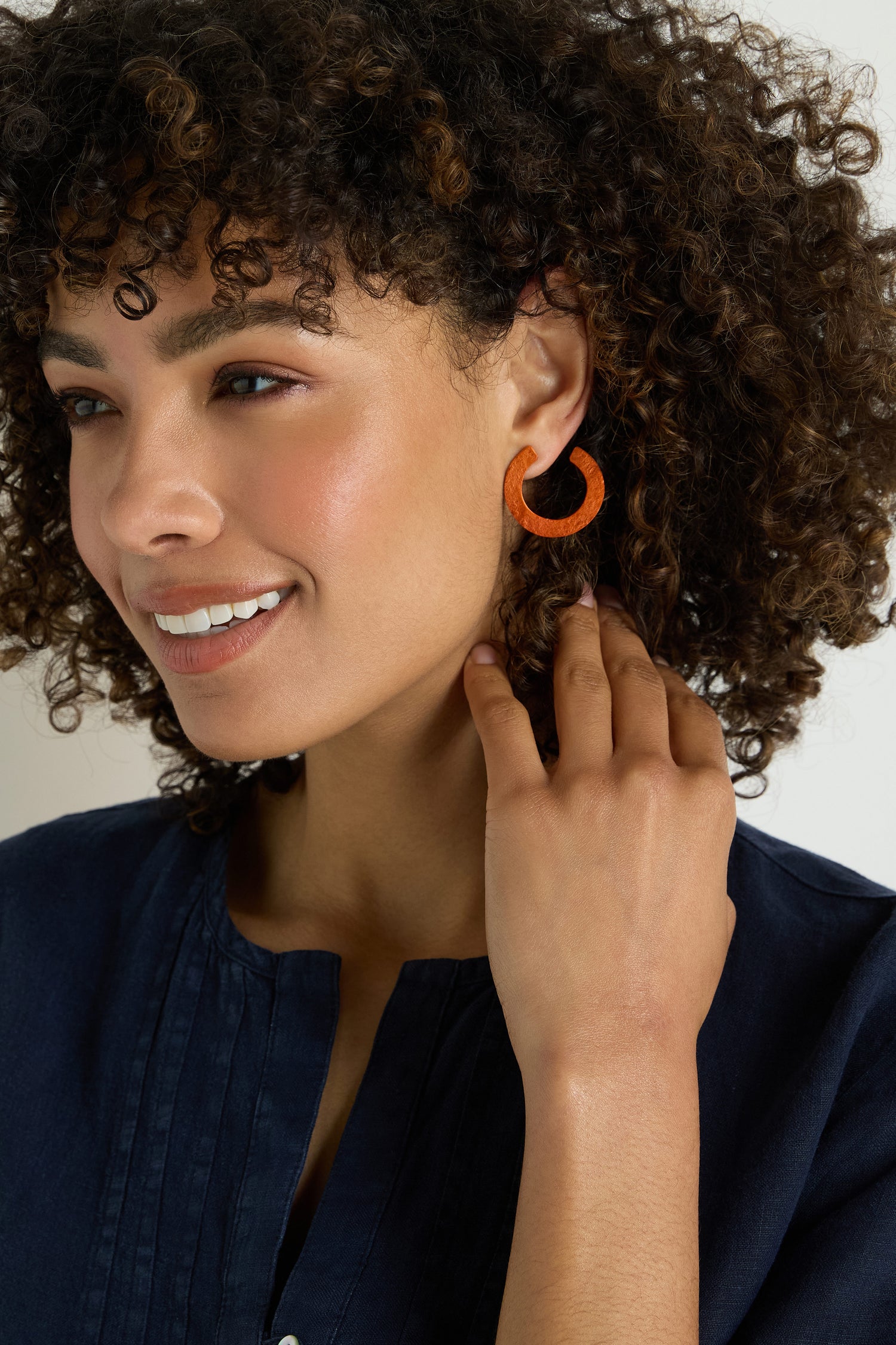 A woman with curly hair, wearing a navy blue top, smiles as she touches her C Curve Earrings.