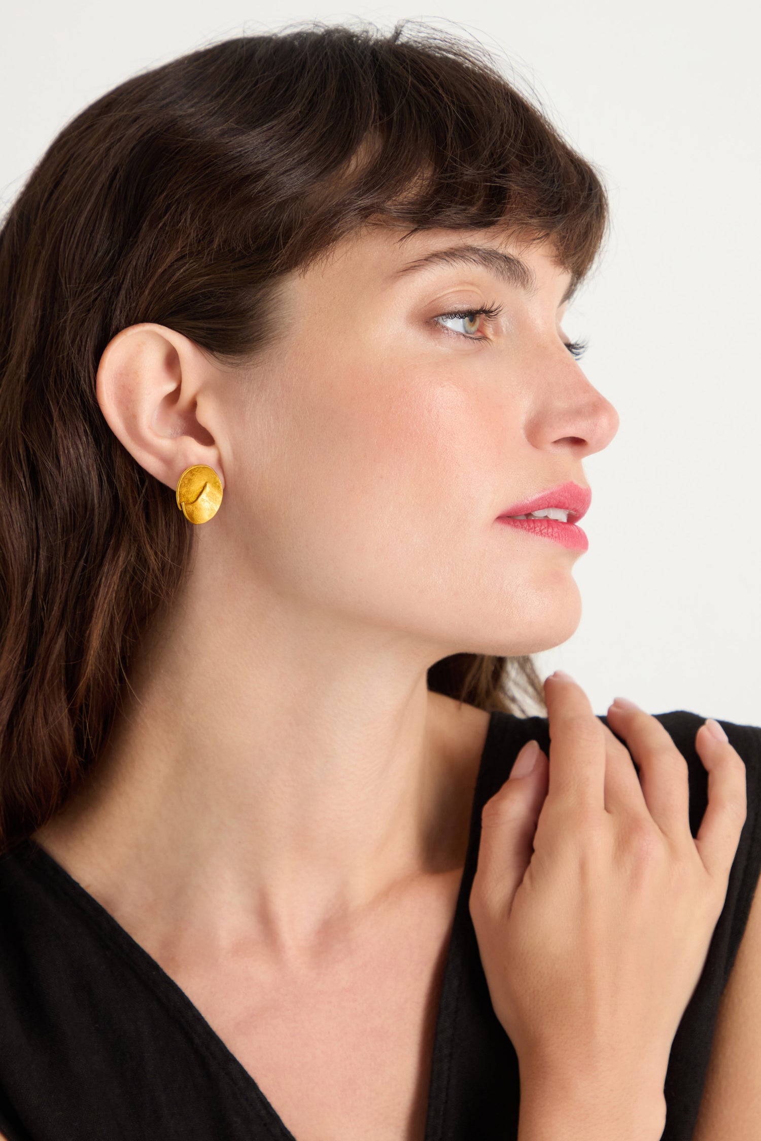 A woman with dark hair wears a black top and Curved Disc Earrings featuring gold texture and sterling silver posts, gazing to the side with her hand near her neck against a plain background.