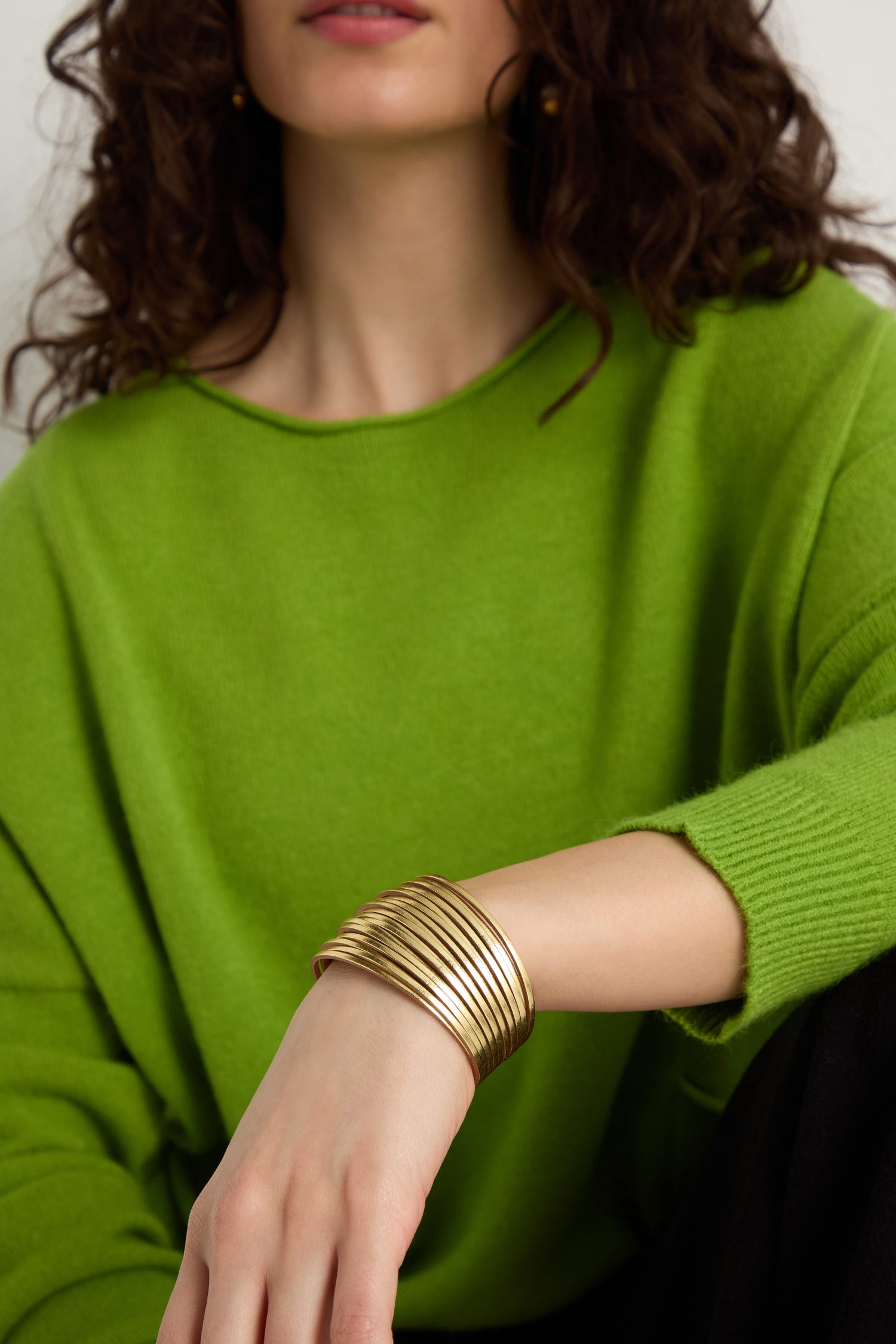 A person wearing a bright green sweater rests their arm on their knee, showing off a Vegan Leather Metallic Bracelet.