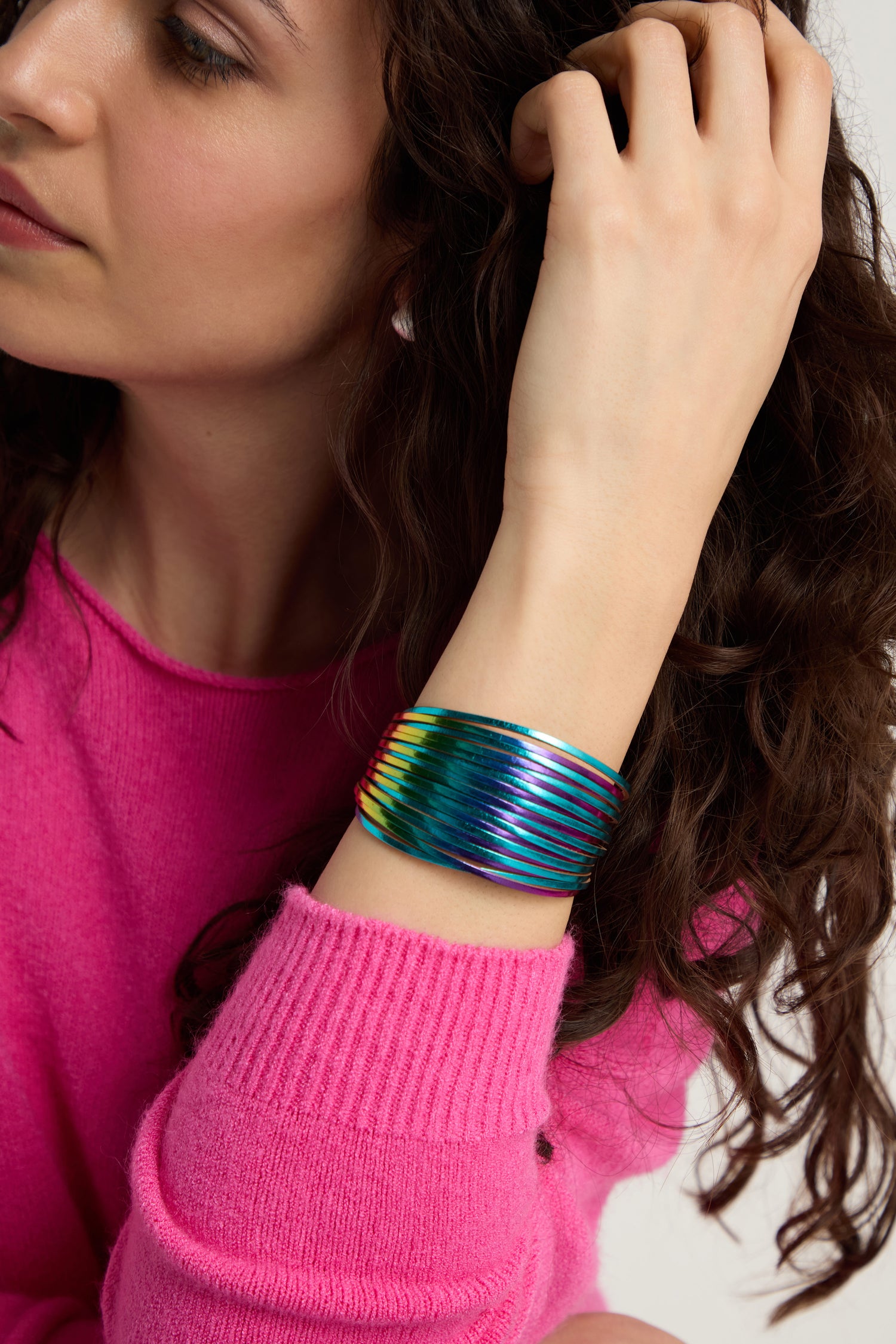 A person wearing a bright pink sweater shows off the Vegan Leather Metallic Bracelet on their wrist while touching their curly brown hair.