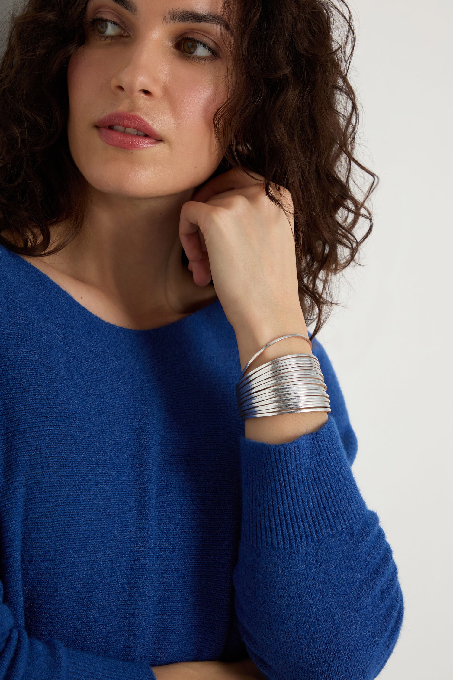 A woman with curly brown hair in a blue sweater rests her hand near her face, wearing the Vegan Leather Metallic Bracelet on her wrist.