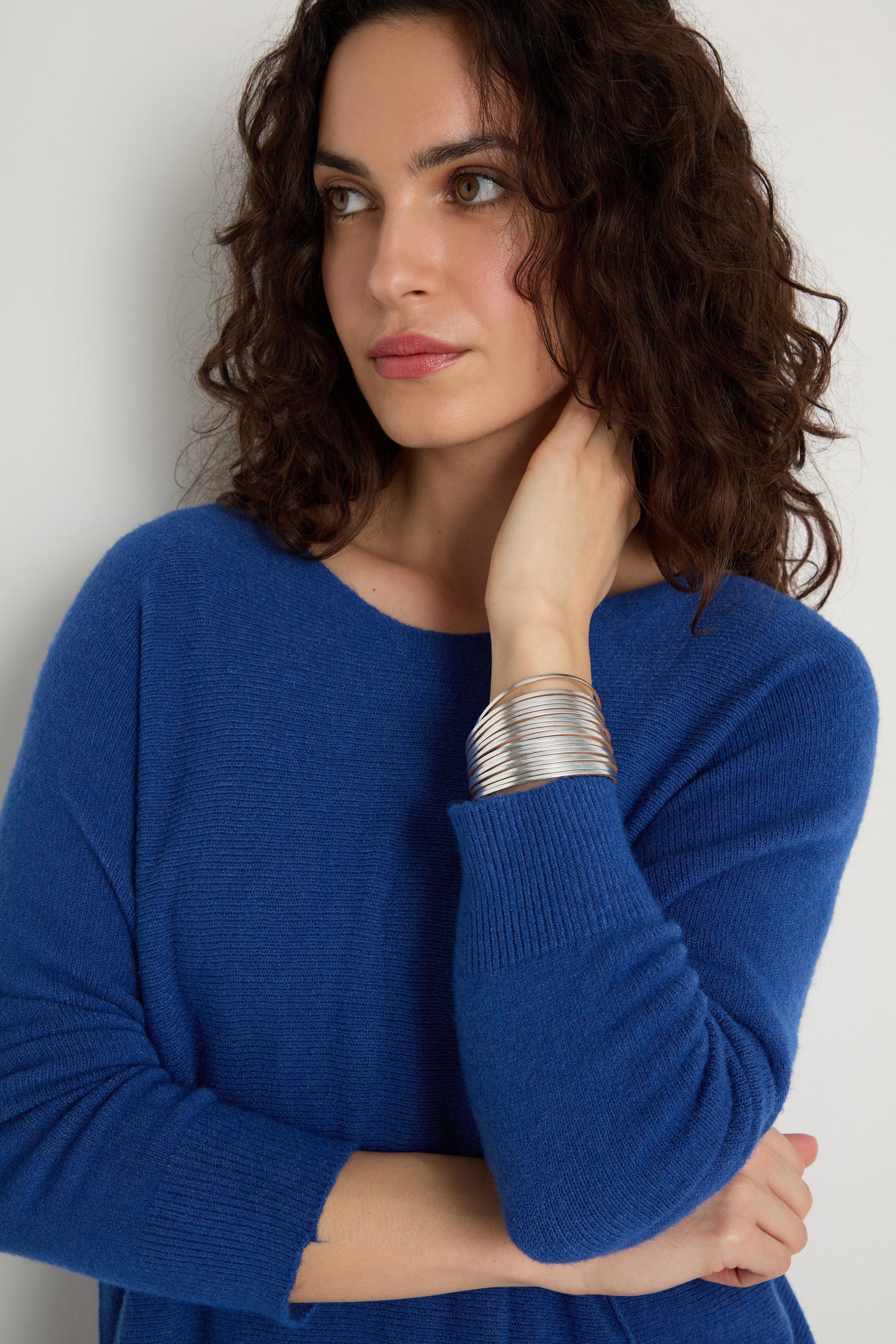 A woman with curly brown hair wears a blue sweater and a Vegan Leather Metallic Bracelet, standing against a white background with one hand touching her neck.