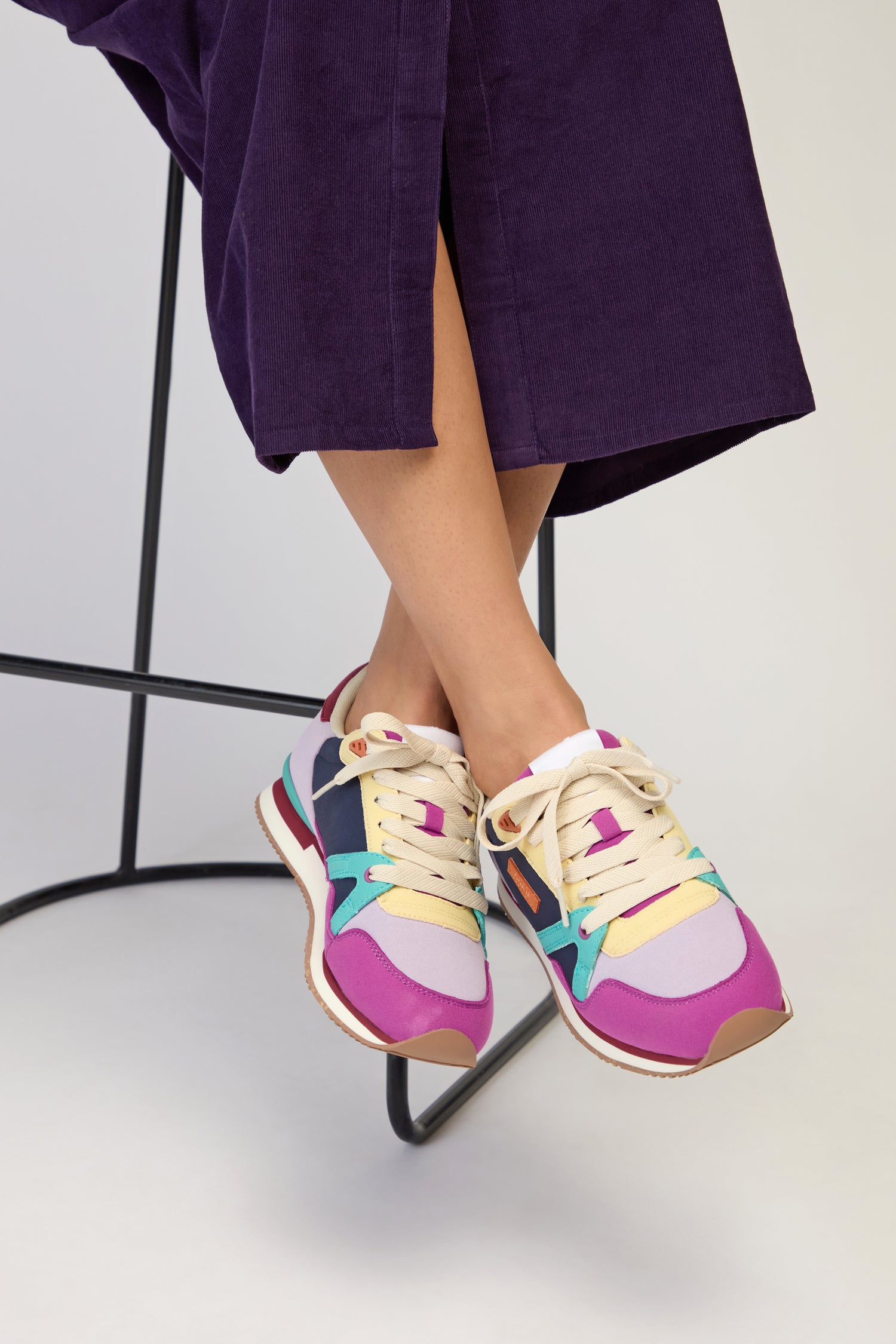 A person wearing a purple skirt and Colour Pop Andree Trainers with bold color blocking sits on a black stool, legs crossed at the ankles.