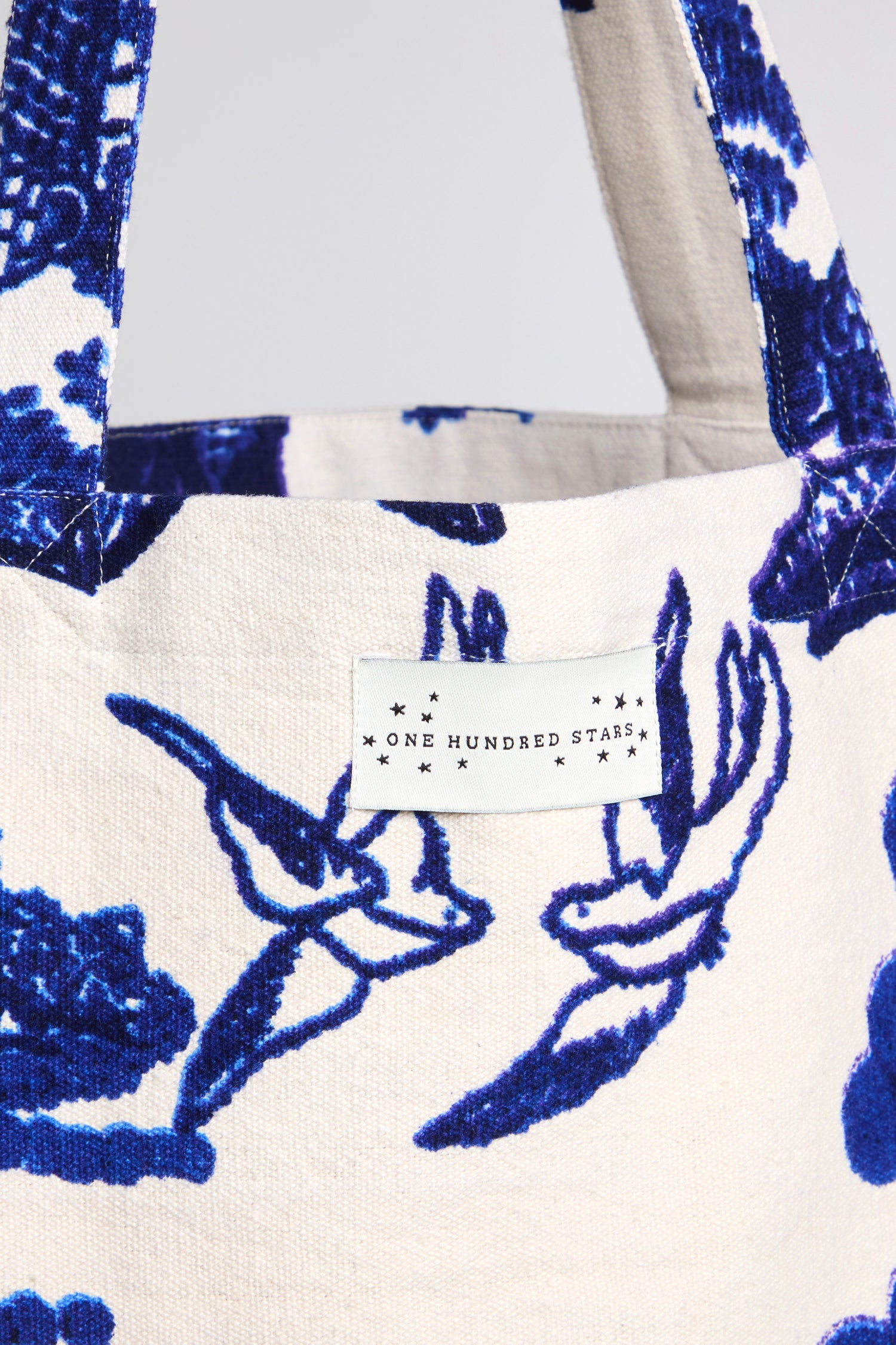 A close-up of the Willow Canvas Bag, showcasing blue birds and floral patterns on white sustainable cotton, with a "One Hundred Stars" label at the top.