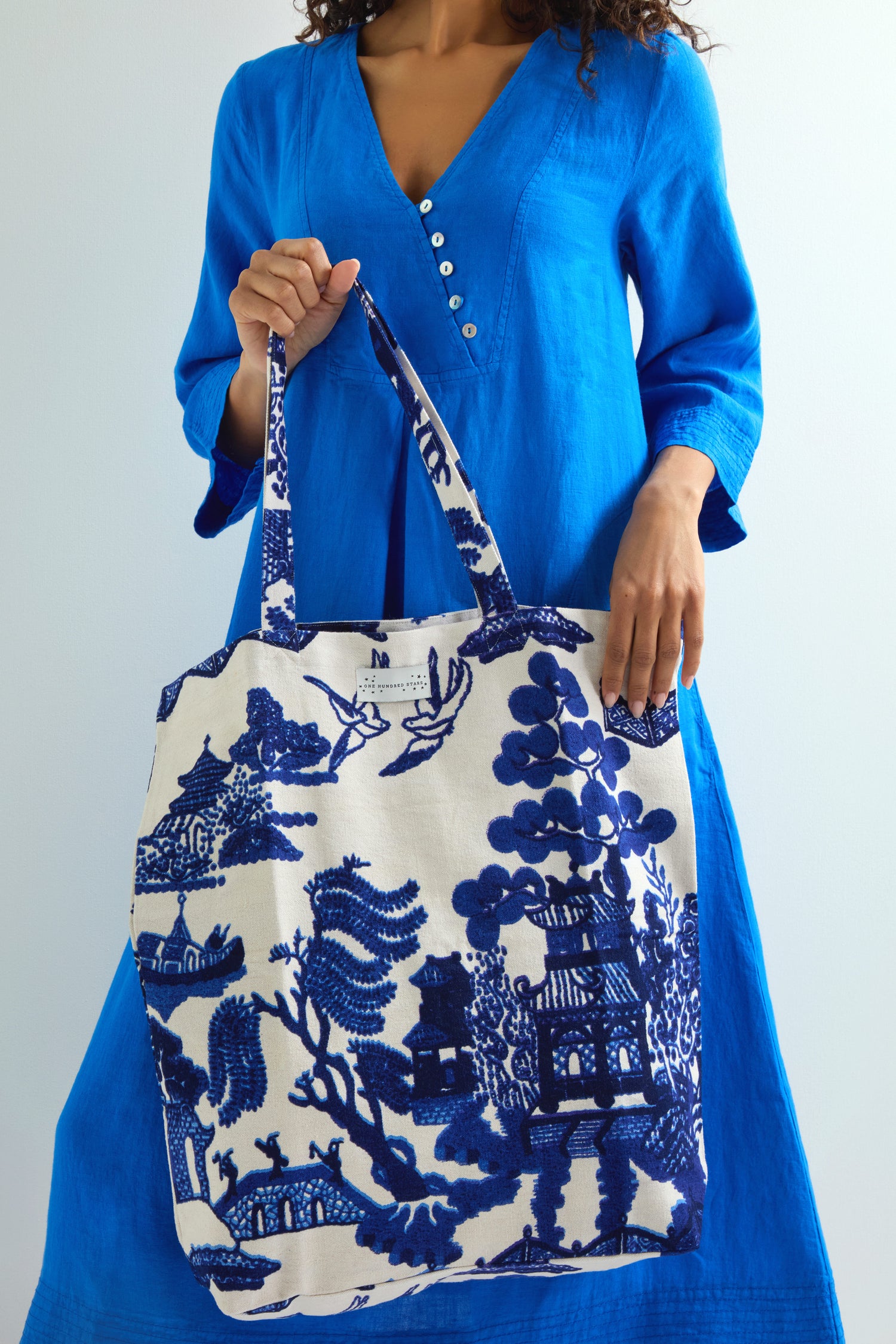 A person in a blue dress holds the Willow Canvas Bag, featuring a blue and white Asian landscape, reminiscent of vintage pottery designs.