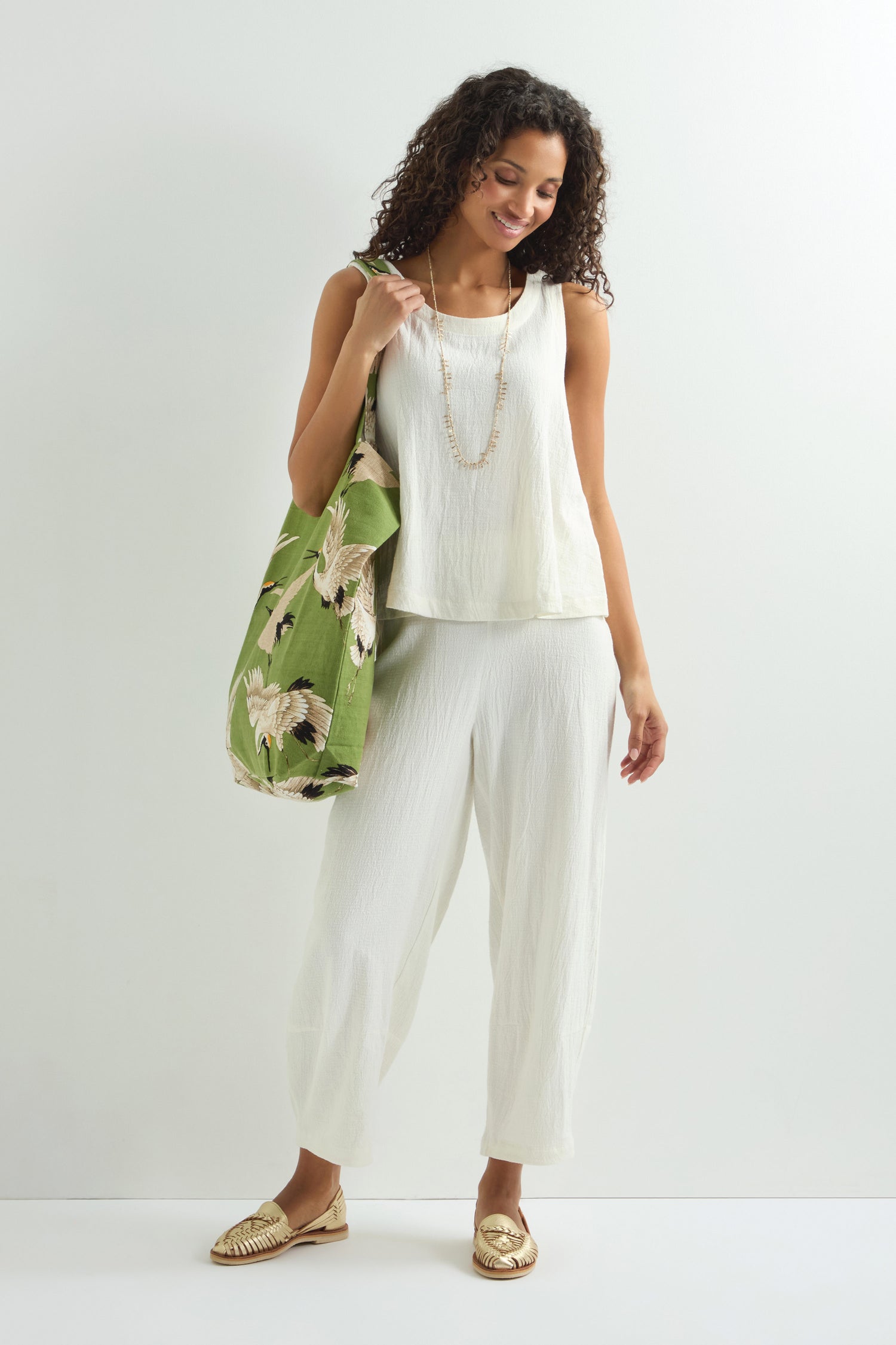 A woman stands against a plain white background, smiling in a white outfit, carrying the Stork Print Canvas Bag with a green floral design over her shoulder. She complements her look with beige woven shoes and a long necklace inspired by Art Deco style.