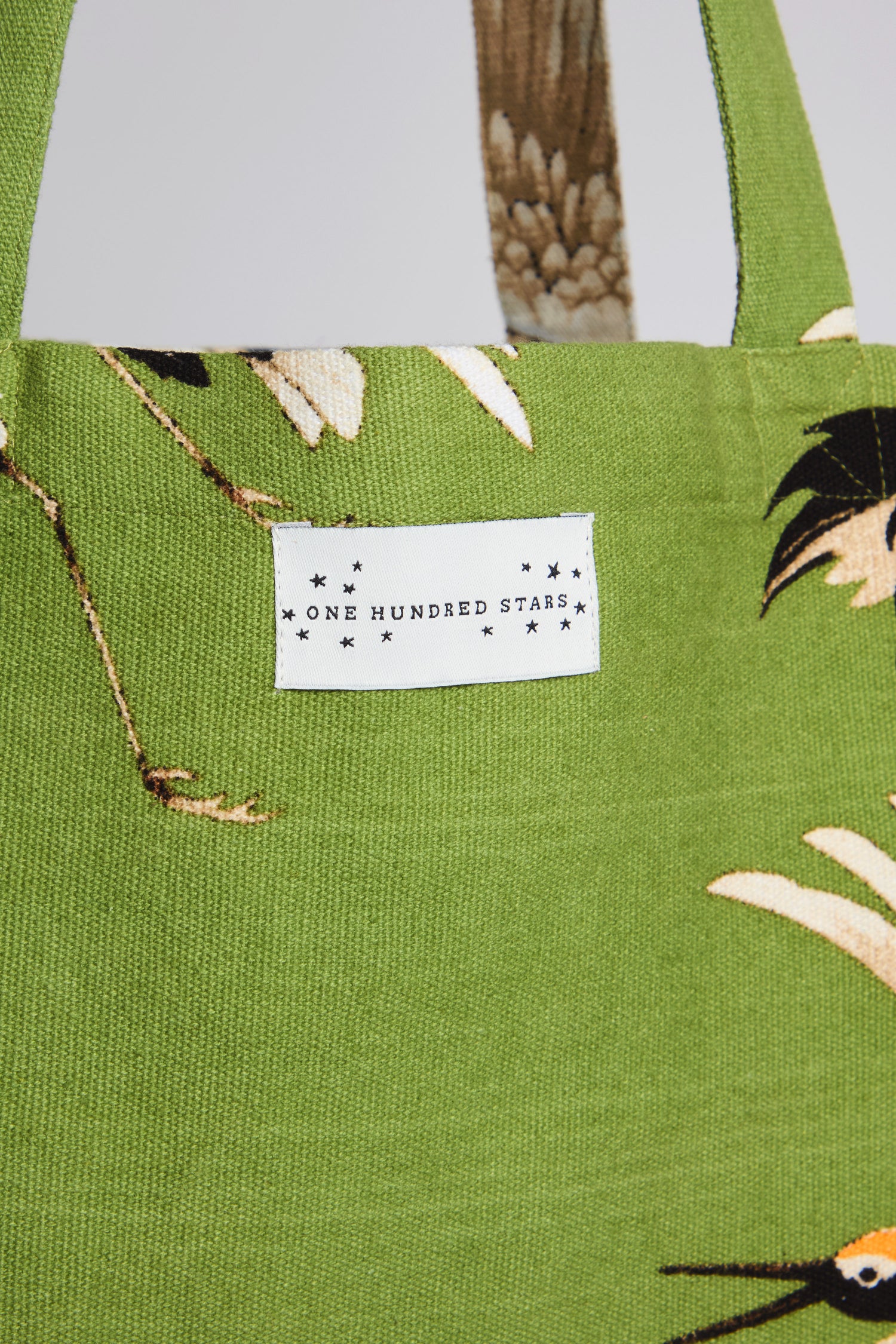 The Stork Print Canvas Bag features a green botanical pattern, showcases a "One Hundred Stars" white tag, and is made from sustainable cotton.