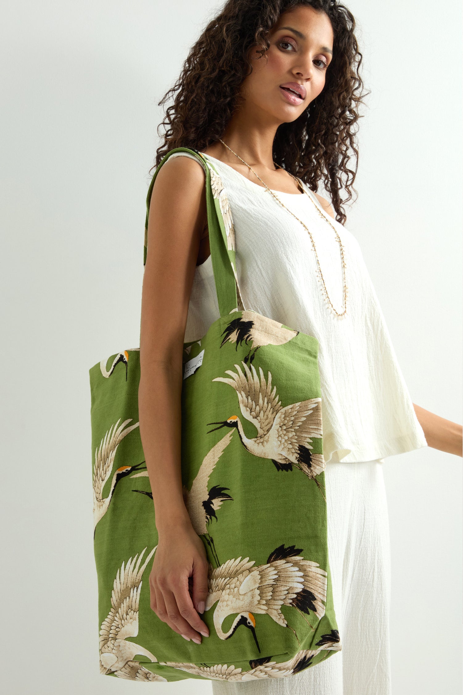 A person in a white outfit holds the Stork Print Canvas Bag, featuring a crane pattern. This large, eco-friendly cotton bag showcases Art Deco-inspired fashion with sustainability.