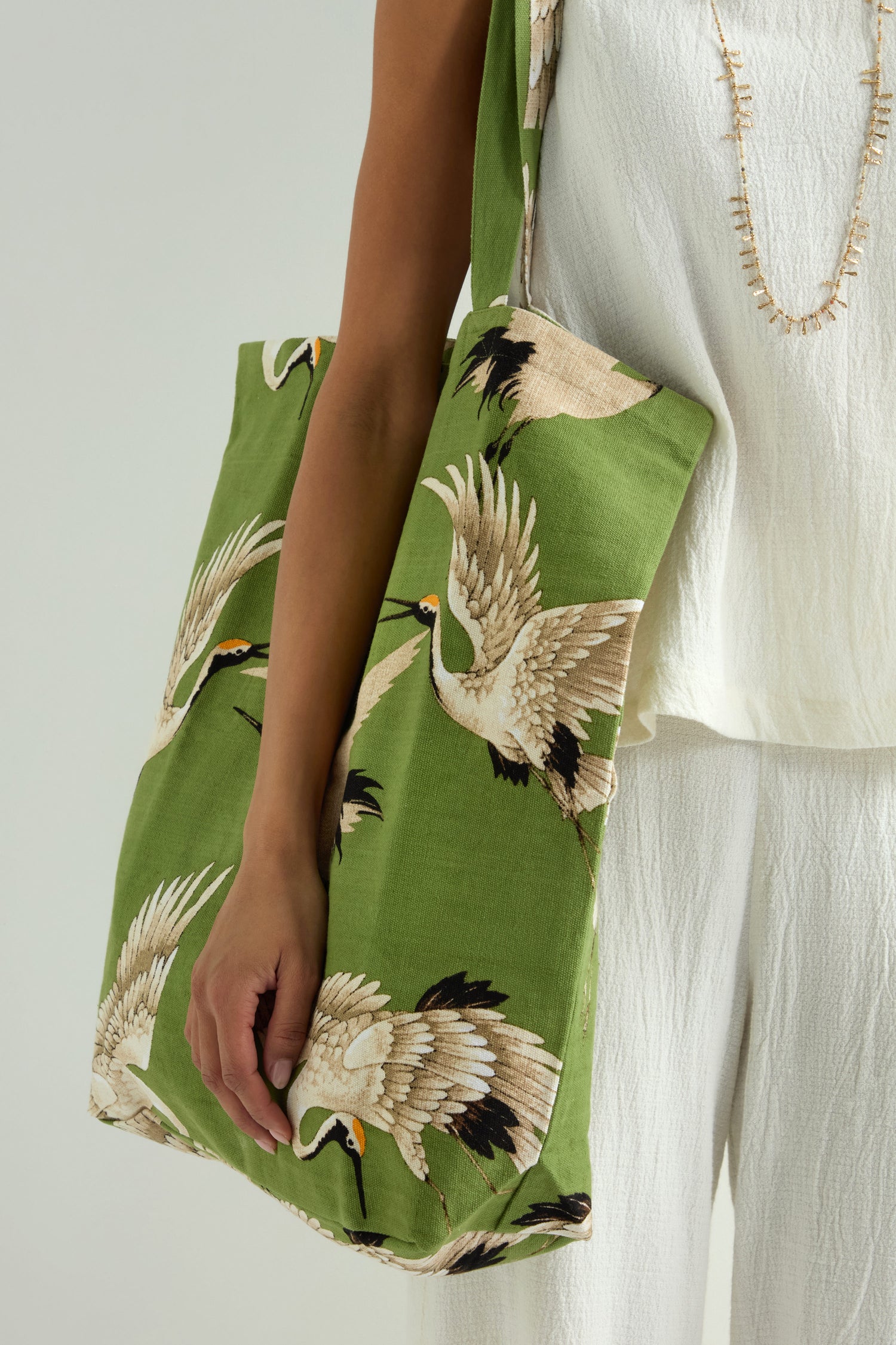A person, dressed in a white outfit and necklace, carries the Stork Print Canvas Bag adorned with crane illustrations, adding an Art Deco touch to their sustainable cotton ensemble.