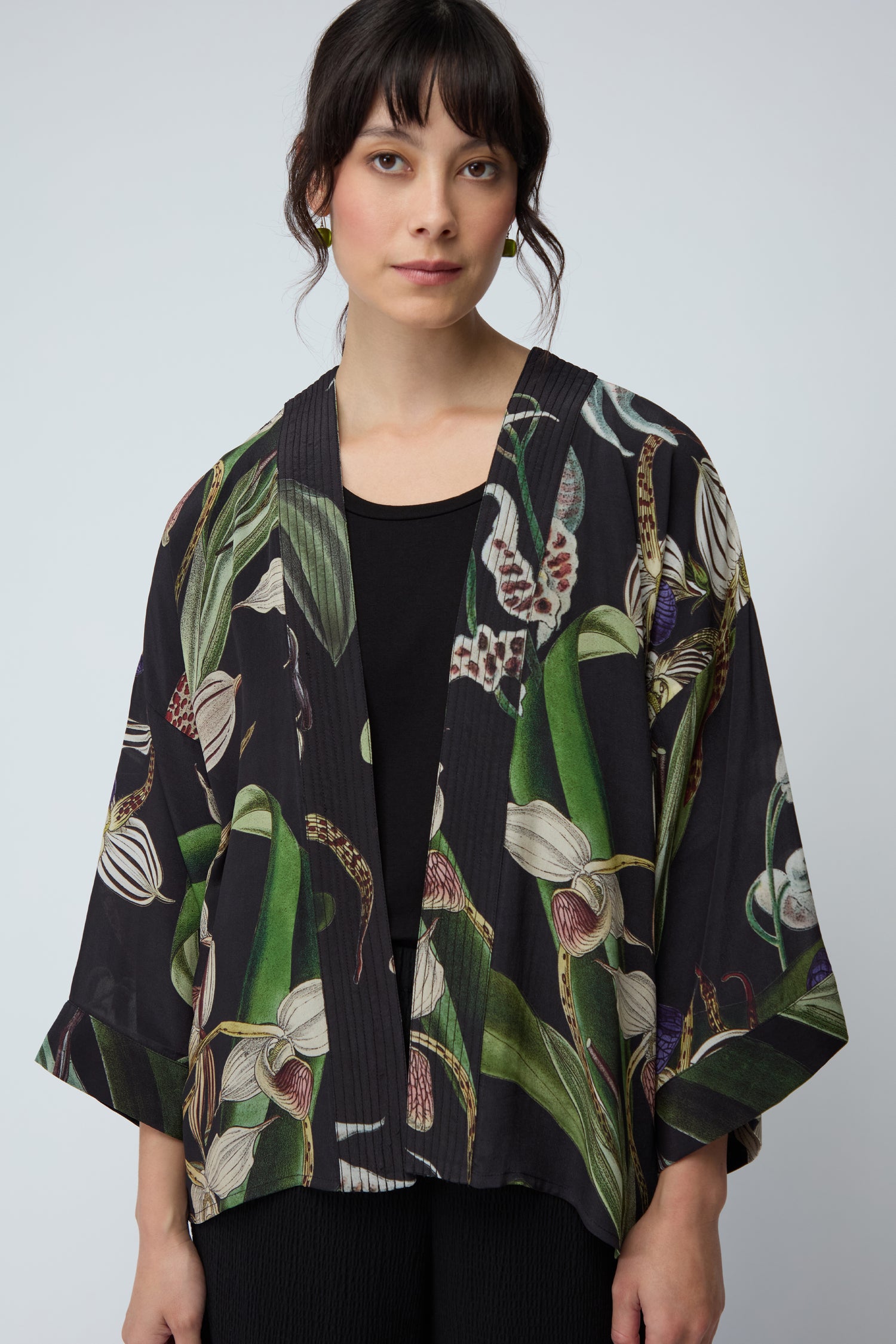 A woman with dark hair wears the Orchid Crepe Kimono, featuring a botanical and butterfly print, over a black top against a plain light background.