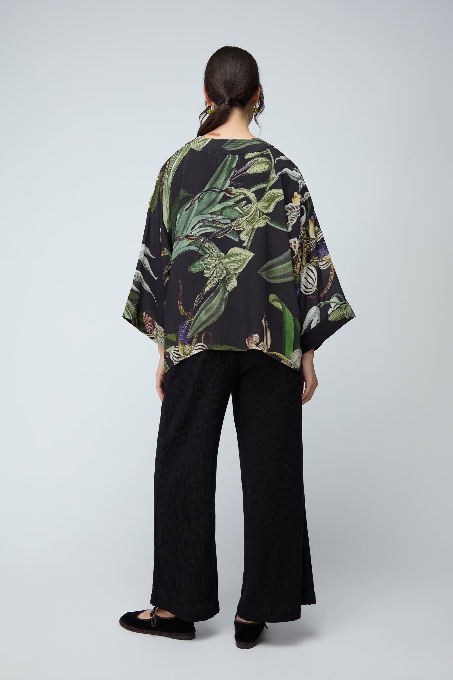 A woman stands facing away, wearing the Orchid Crepe Kimono in a black botanical print with sustainable fabric, paired with black wide-leg pants and shoes, set against a plain light background.