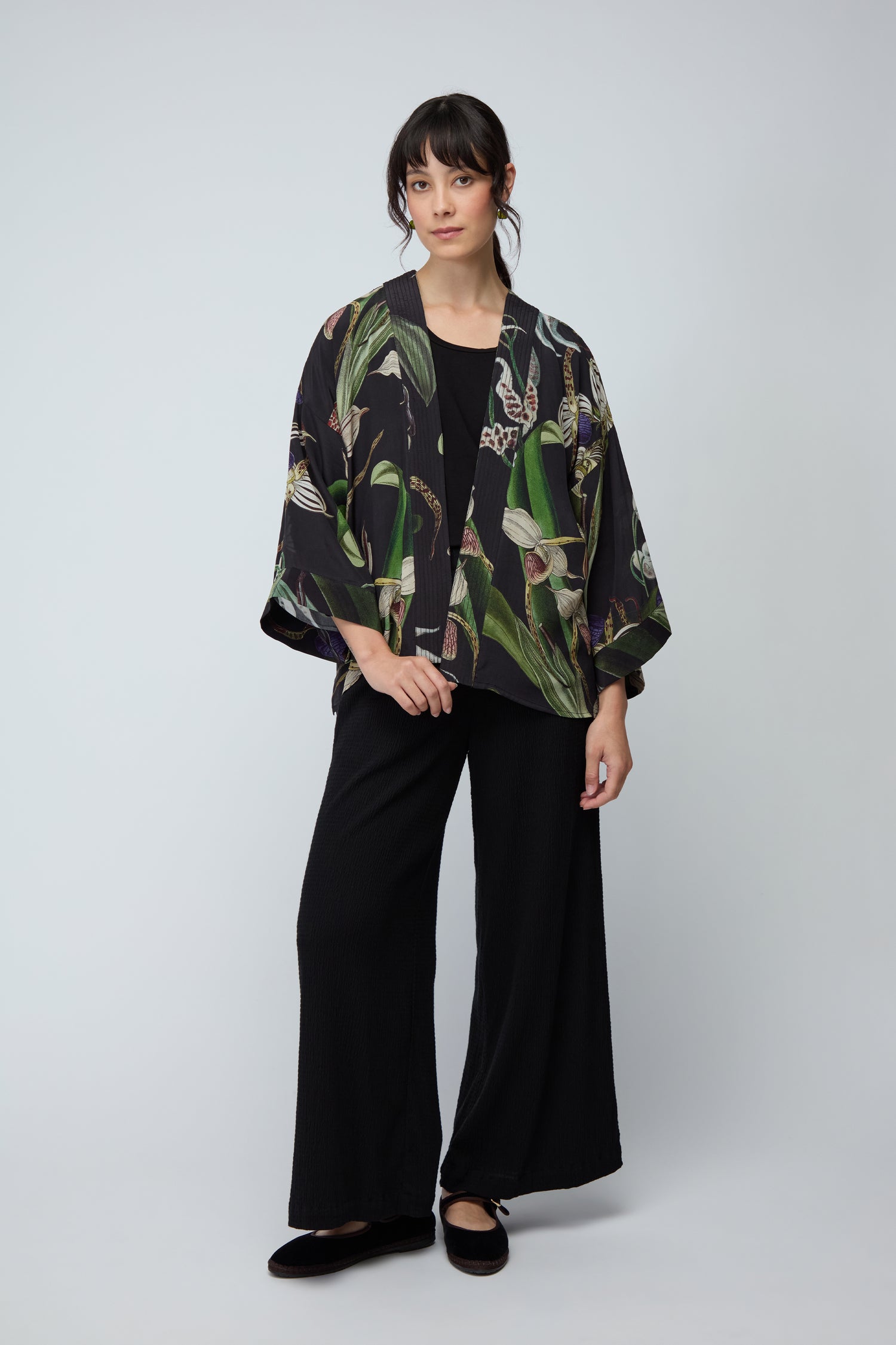 A woman stands against a plain background wearing the Orchid Crepe Kimono with a black botanical print, made from sustainable fabric, paired with black wide-leg pants and black shoes.