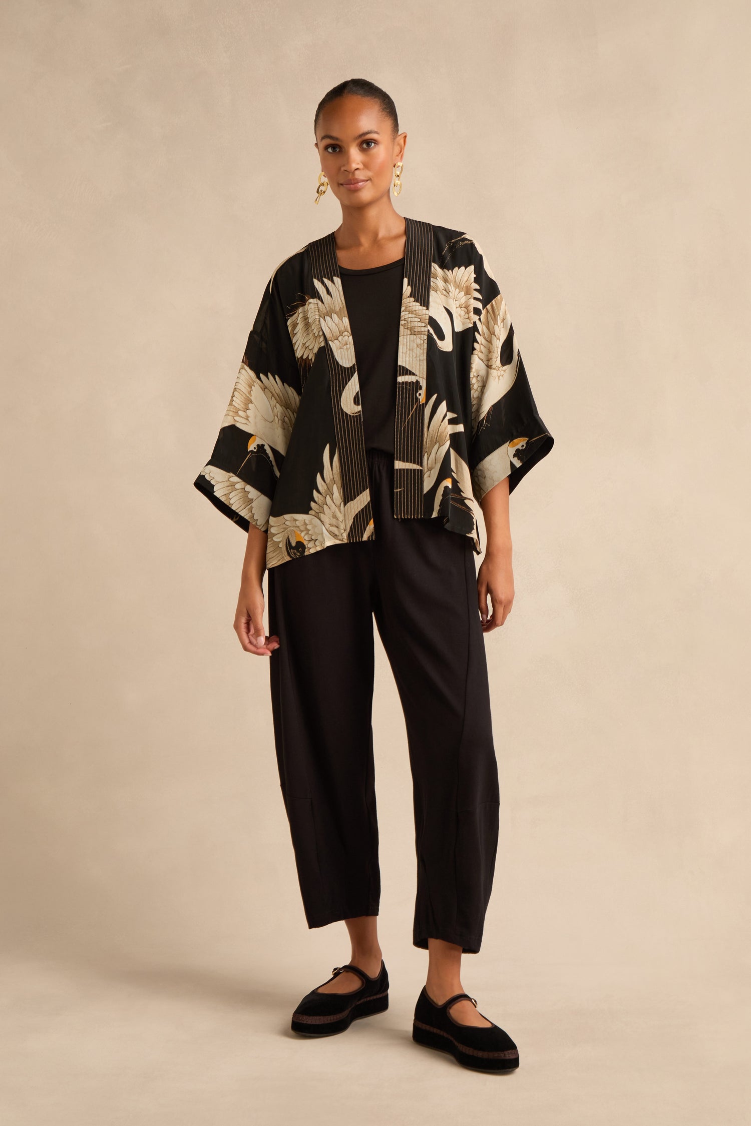 Person standing against a plain background wearing black pants, black shoes, a black top, and the Stork Crepe Kimono featuring an elegant crane bird motif inspired by Art Deco design.