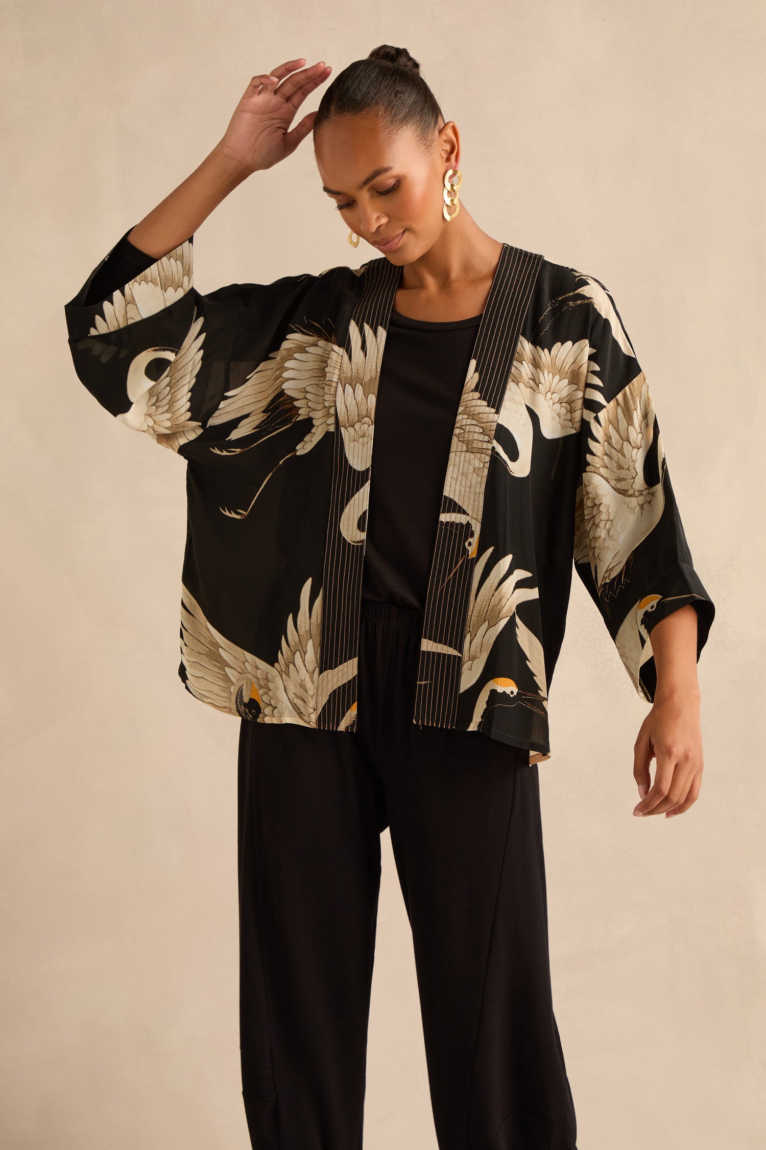 A woman stands against a beige background wearing a black outfit layered with the Stork Crepe Kimono, which showcases an exclusive Art Deco stork print.