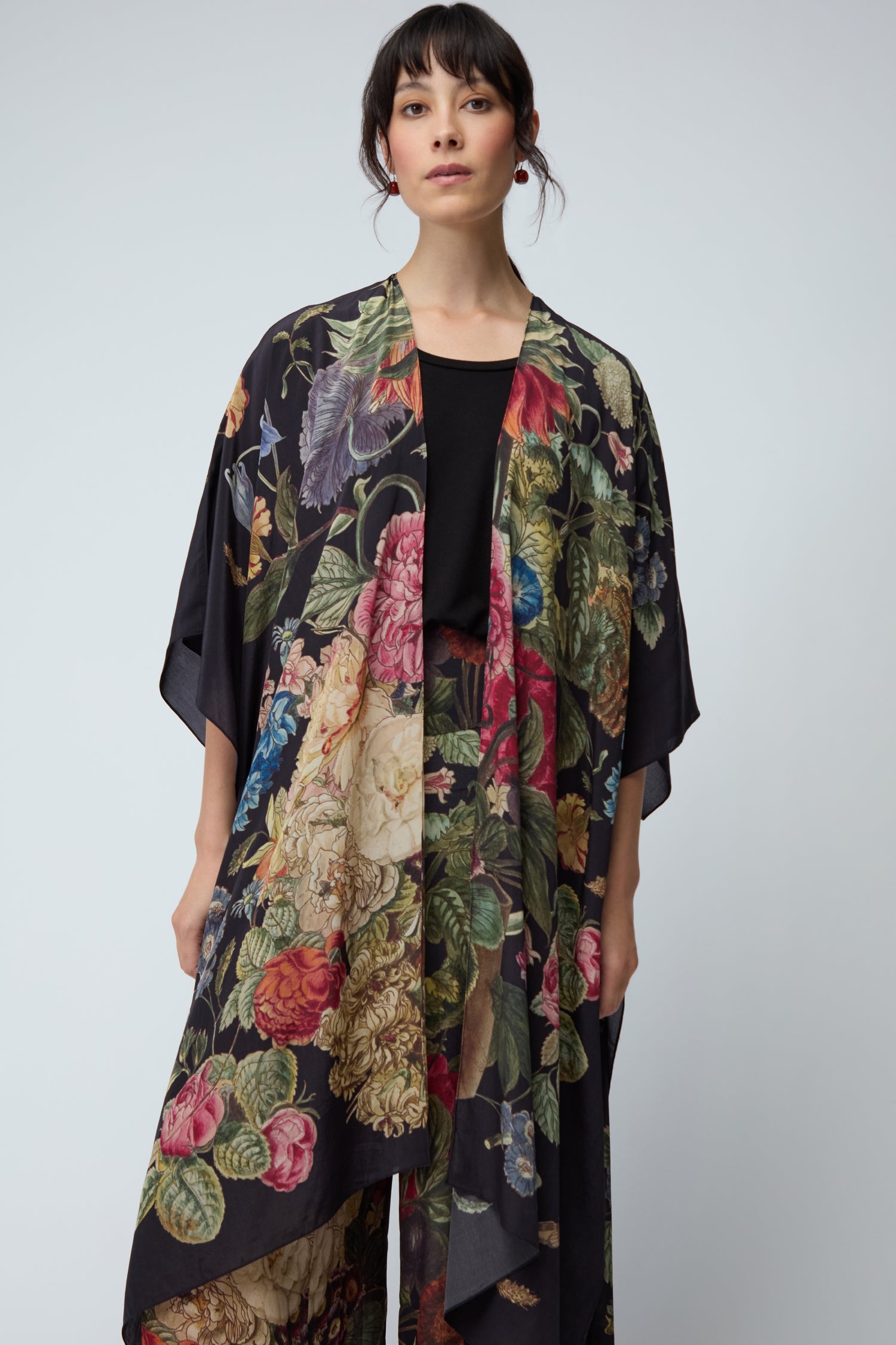 A woman wears the Primula Crepe Throwover, a black kimono-style cover-up with a vibrant floral pattern, layered over a black top and standing against a plain light background.
