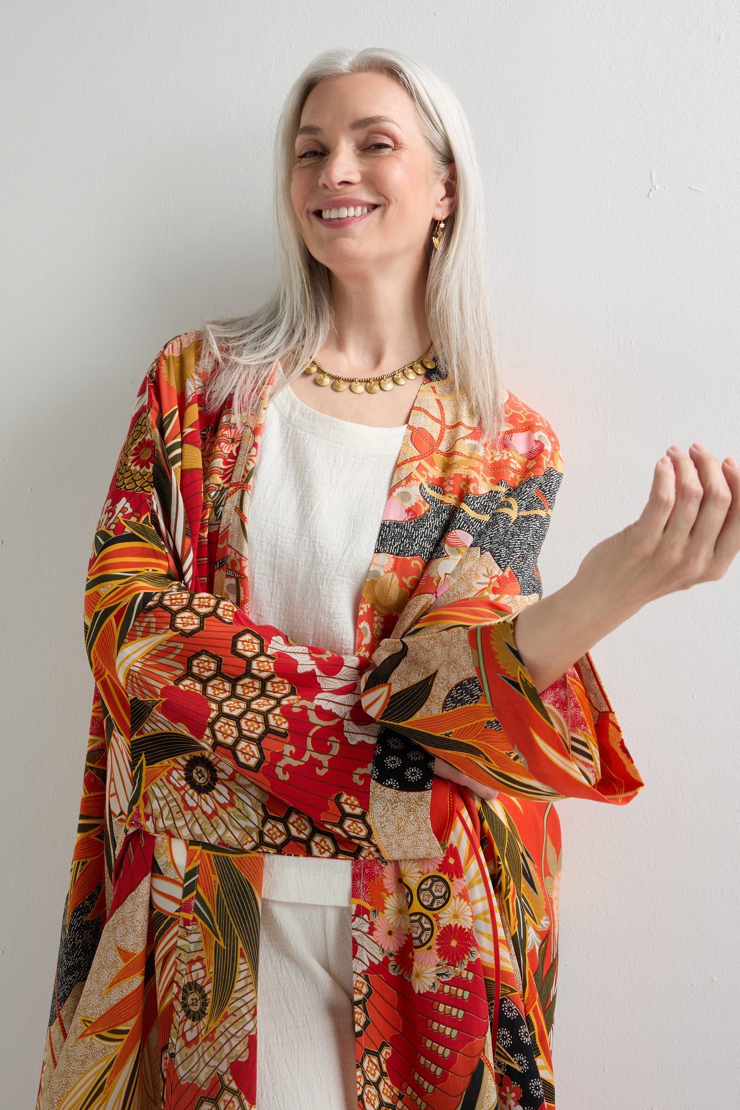 A woman with long gray hair in a white outfit and the Fans Print Grande Kimono smiles against a plain white wall, highlighting the traditional artistry of this standout layer.