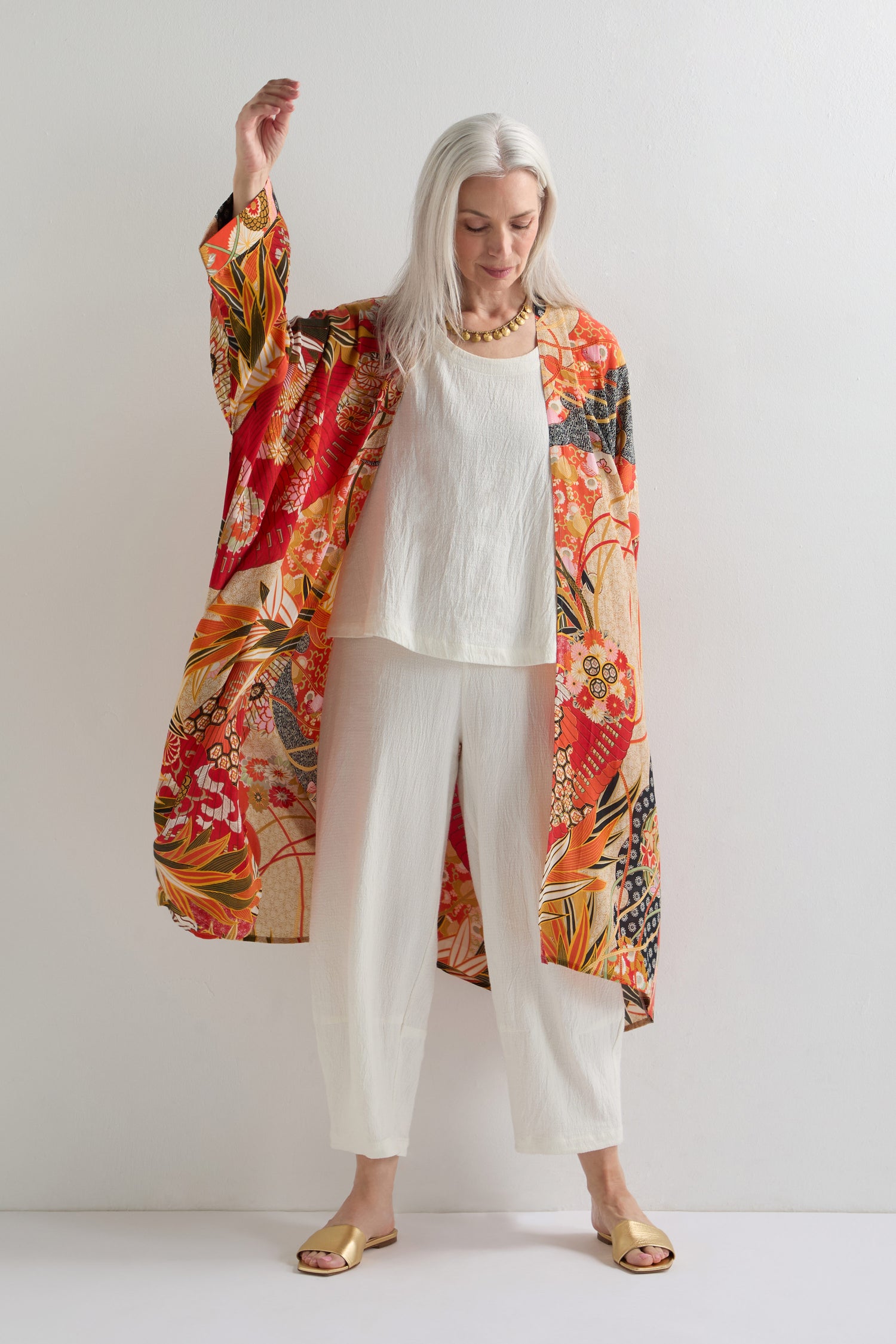 A woman with long gray hair stands against a white wall, wearing the Fans Print Grande Kimono over a white outfit with gold sandals, highlighting its statement style and traditional artistry.