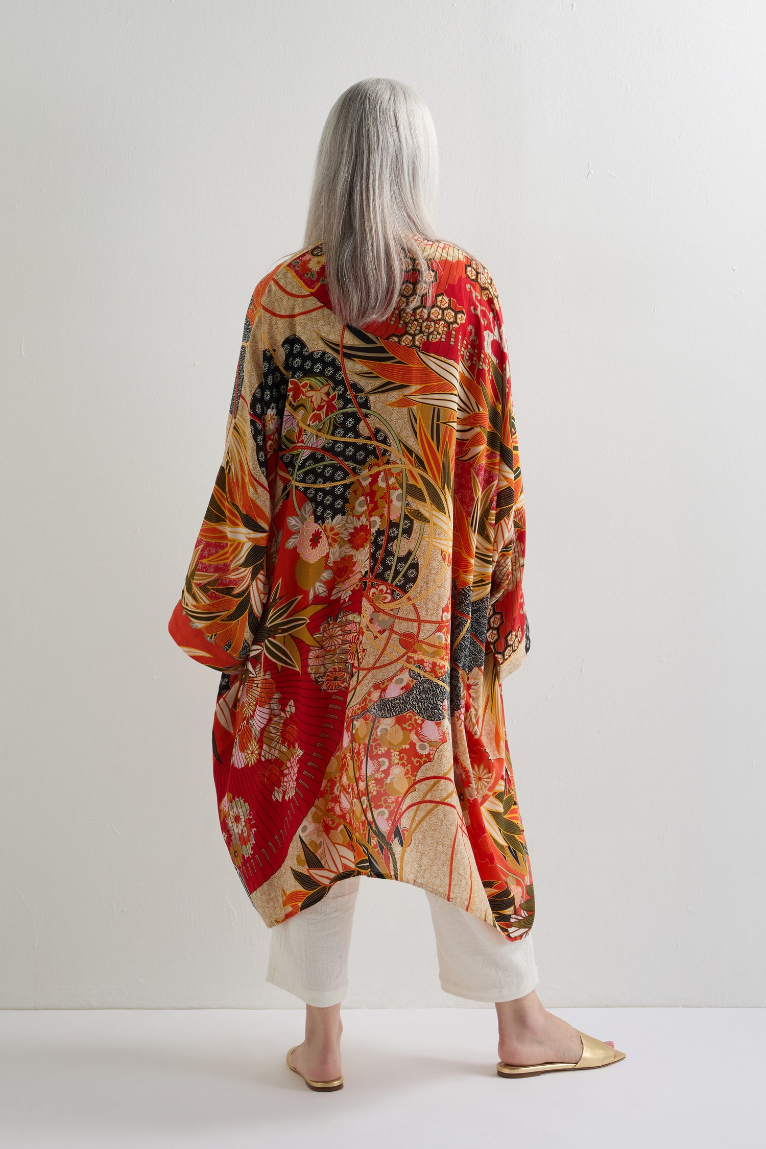A person with long gray hair stands barefoot, facing a white wall and wearing the Fans Print Grande Kimono in red and gold over white pants, making a bold statement with its traditional artistry.