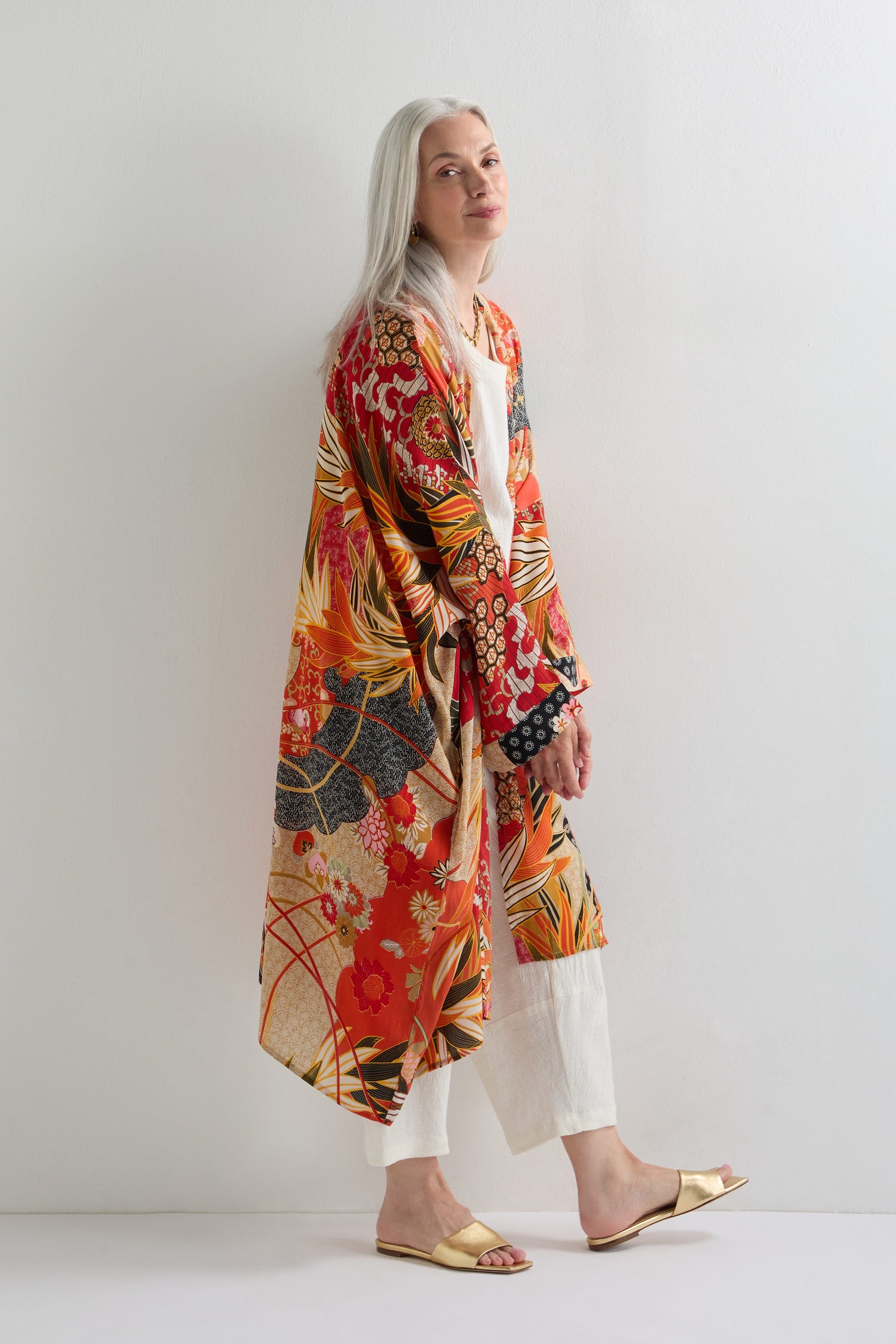 A woman with long gray hair wears the Fans Print Grande Kimono over a white outfit and sandals, standing against a plain white background.