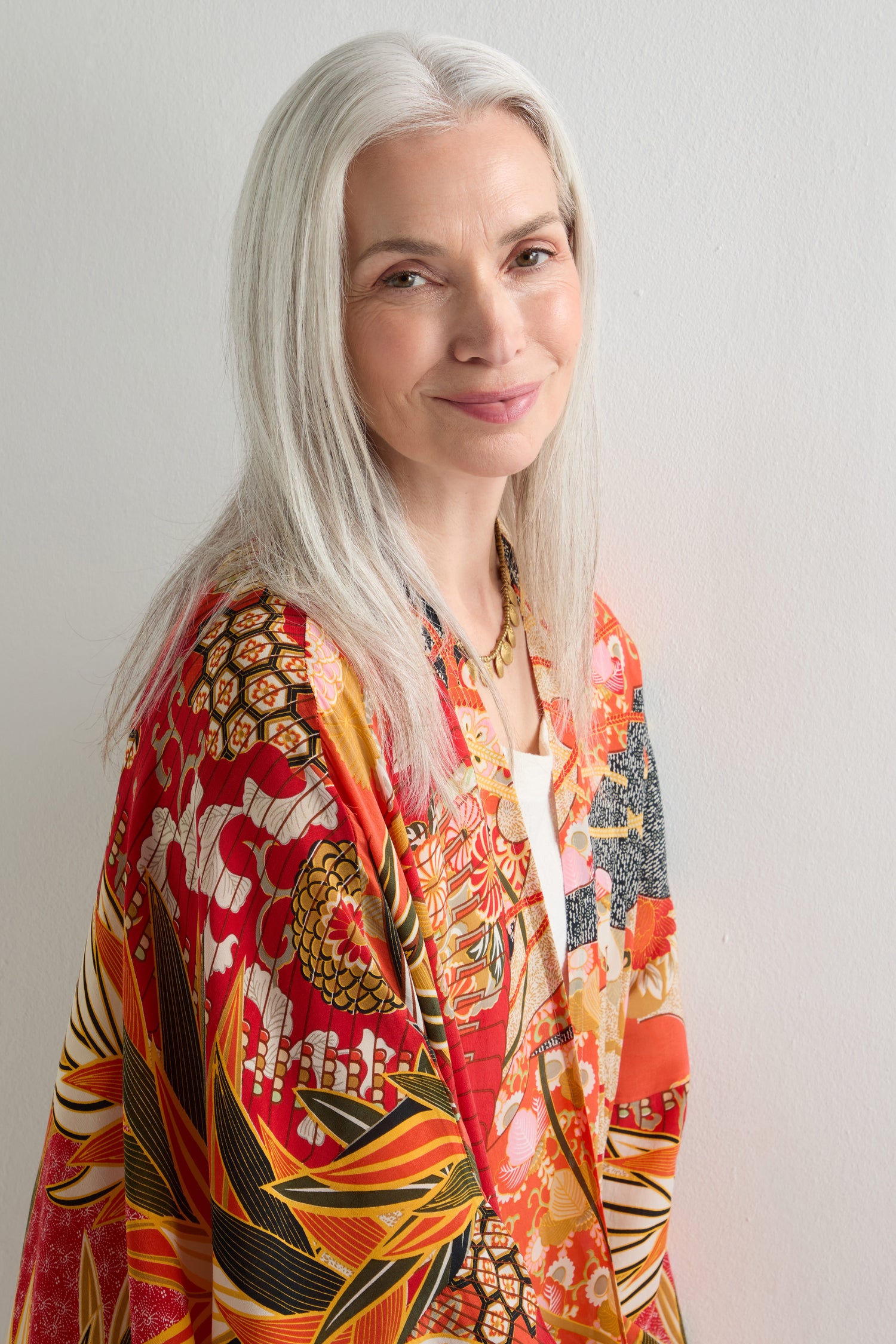 An older woman with long gray hair smiles softly in the Fans Print Grande Kimono, its colorful design celebrating traditional artistry against a plain white wall.