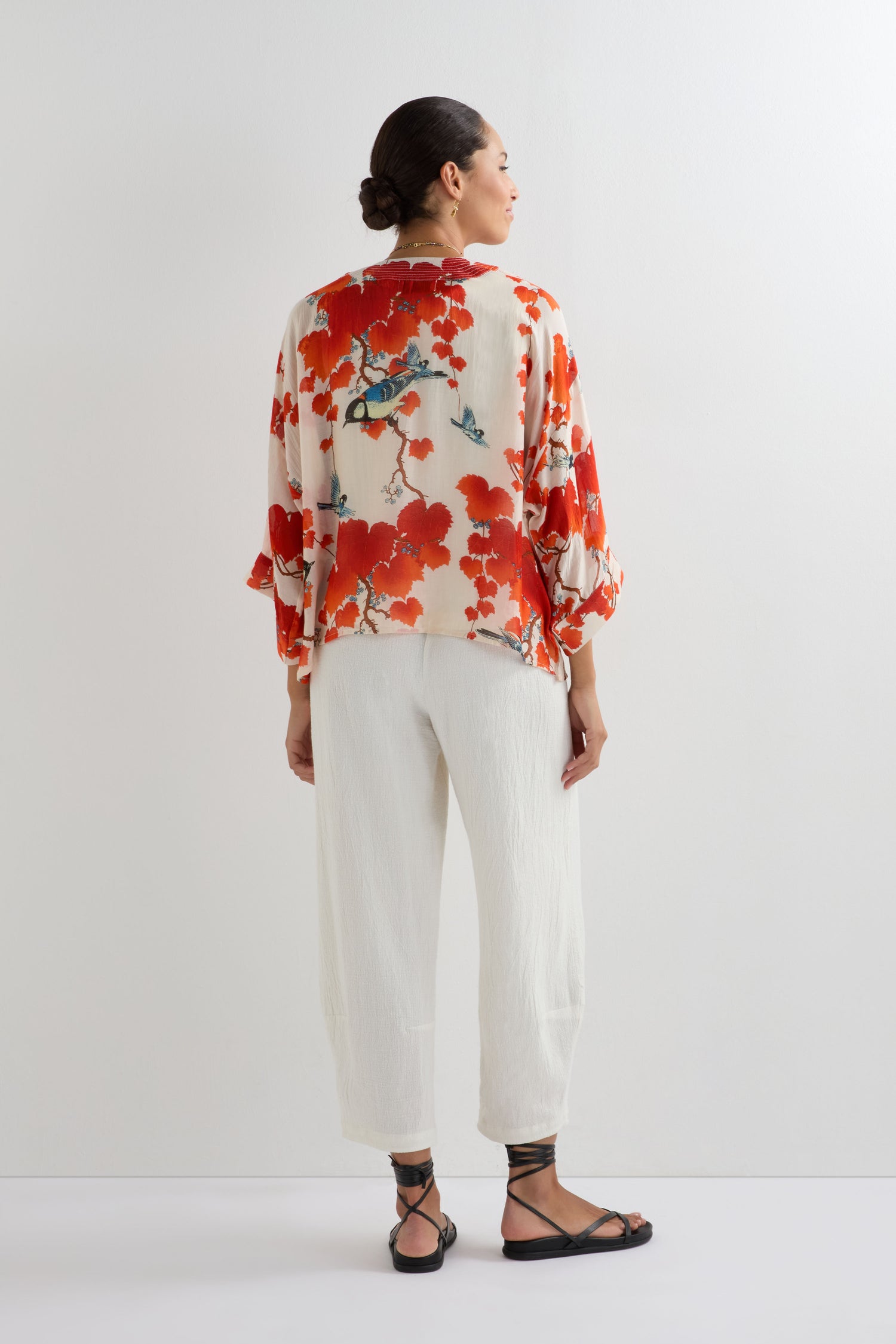 A person stands facing away, wearing the Acer Print Kimono with red summer-inspired floral patterns, paired with white pants and black strappy sandals against a plain white background.