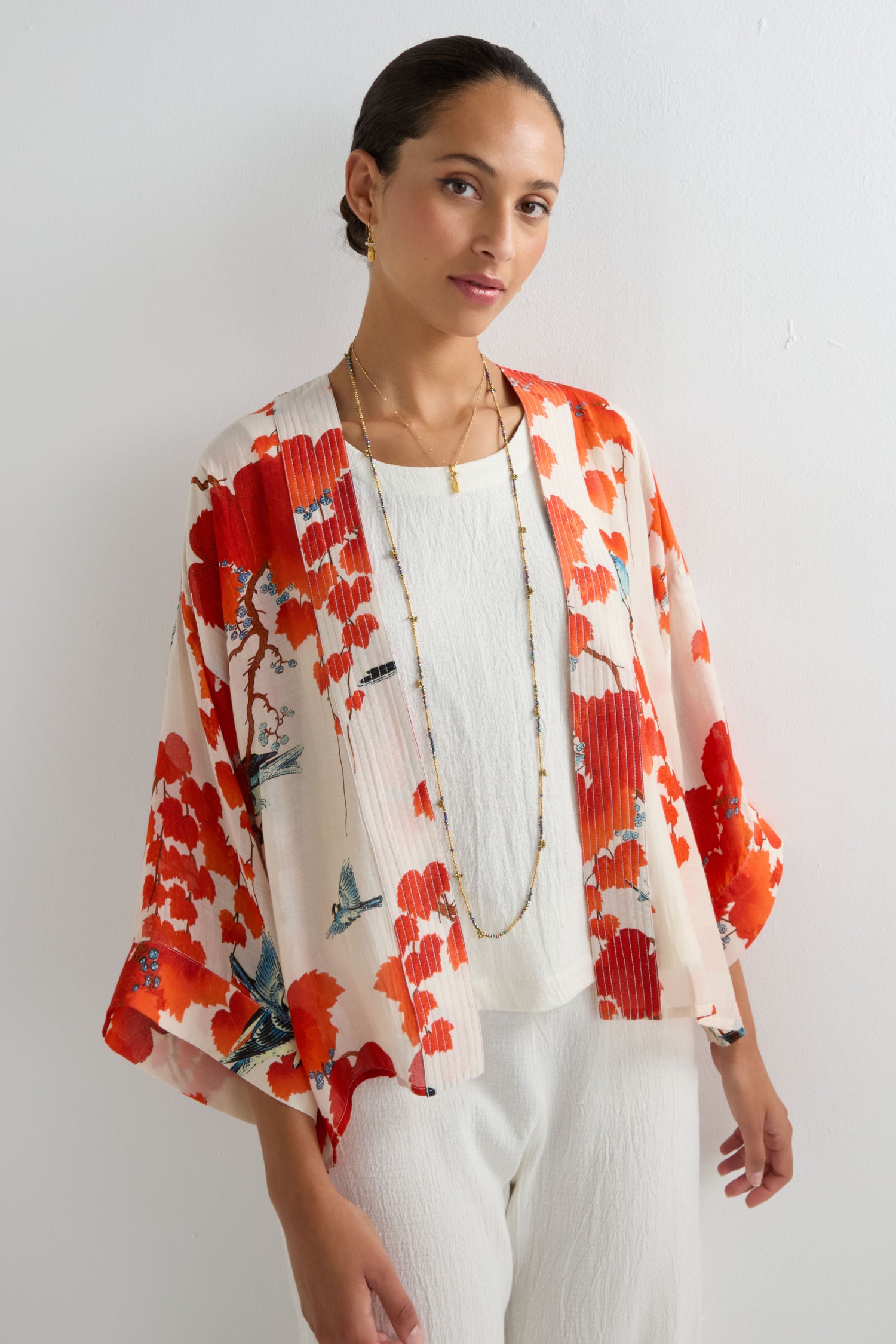 A woman stands against a white wall wearing a white outfit, layered necklaces, and the Acer Print Kimono featuring vibrant red and orange prints inspired by Japanese maple leaves.