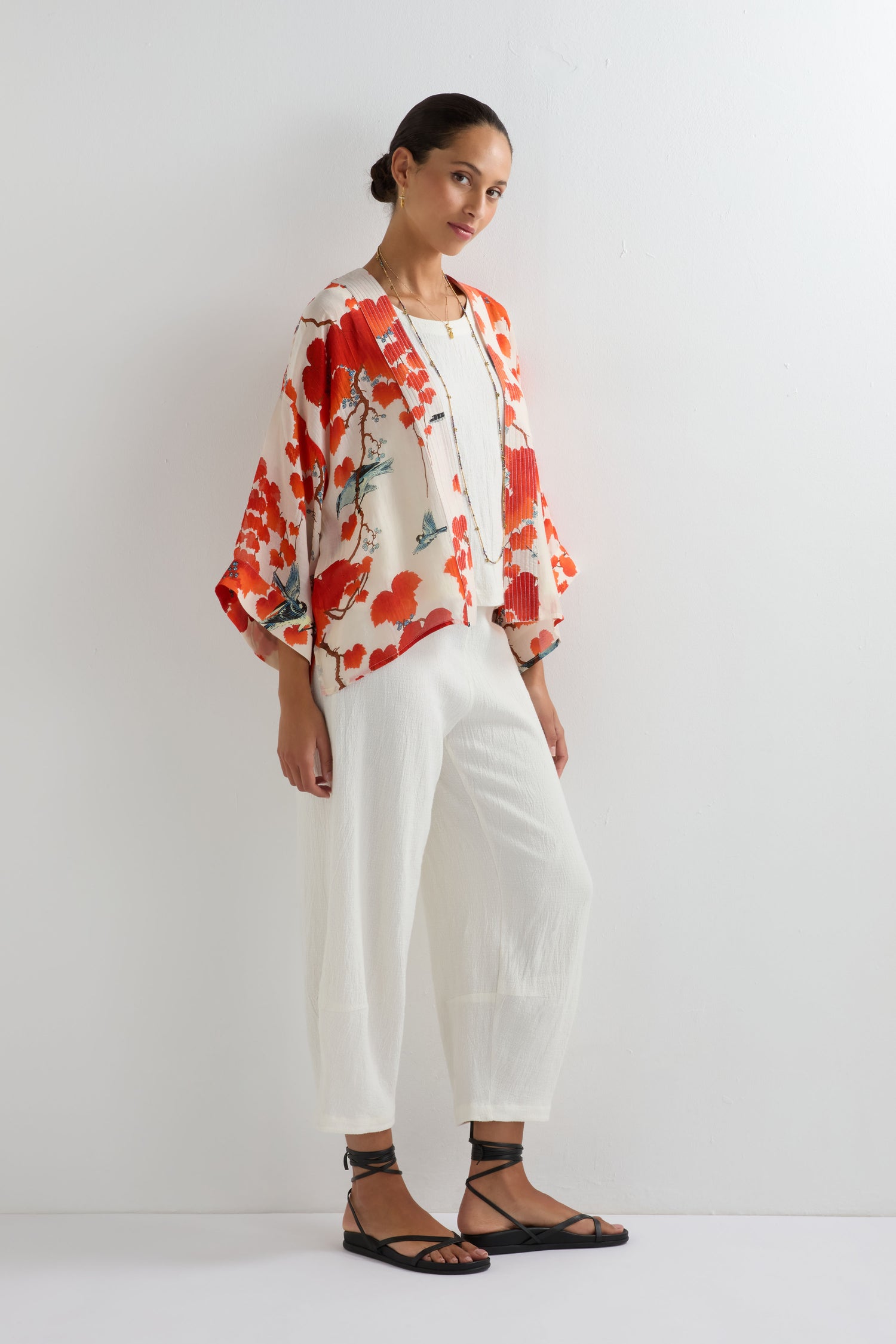 A woman stands against a white wall wearing a white jumpsuit, black sandals, and the Acer Print Kimono in floral red and cream, inspired by summer prints.
