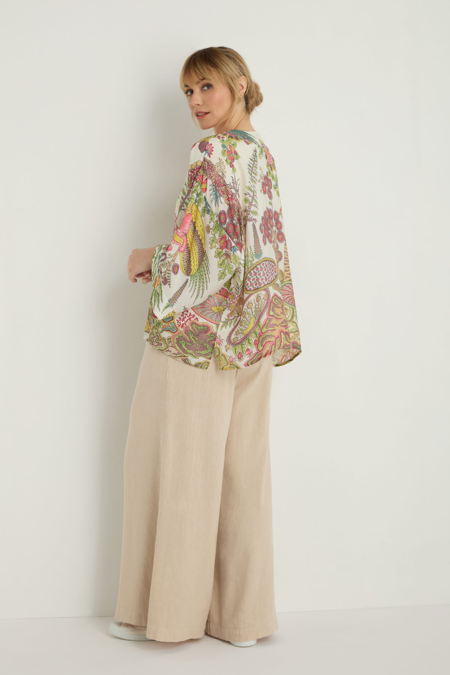 A woman stands against a white wall, facing slightly away and looking back. She wears the Boho Garden Ecru Print Kimono over loose, beige wide-leg pants, showcasing its lightweight feel and vintage-inspired motifs.