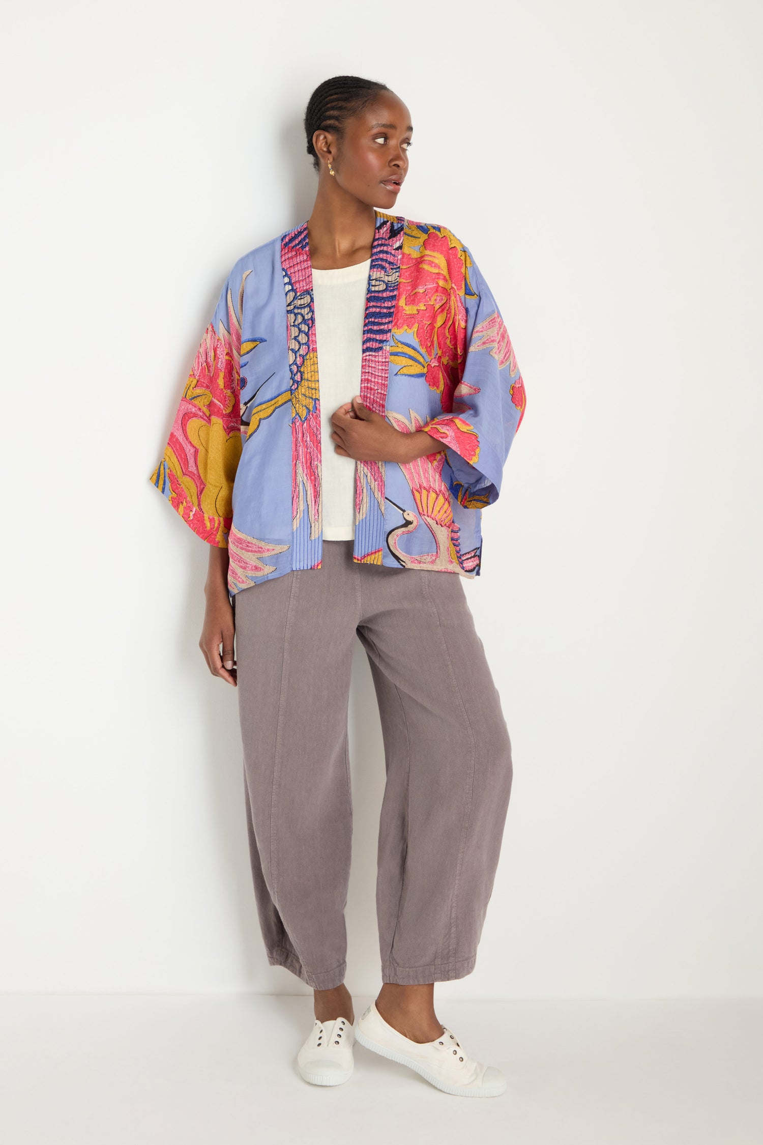 A person stands against a white wall wearing an eye-catching Crane Slate Print Kimono, a light beige top, loose gray pants, and white sneakers.