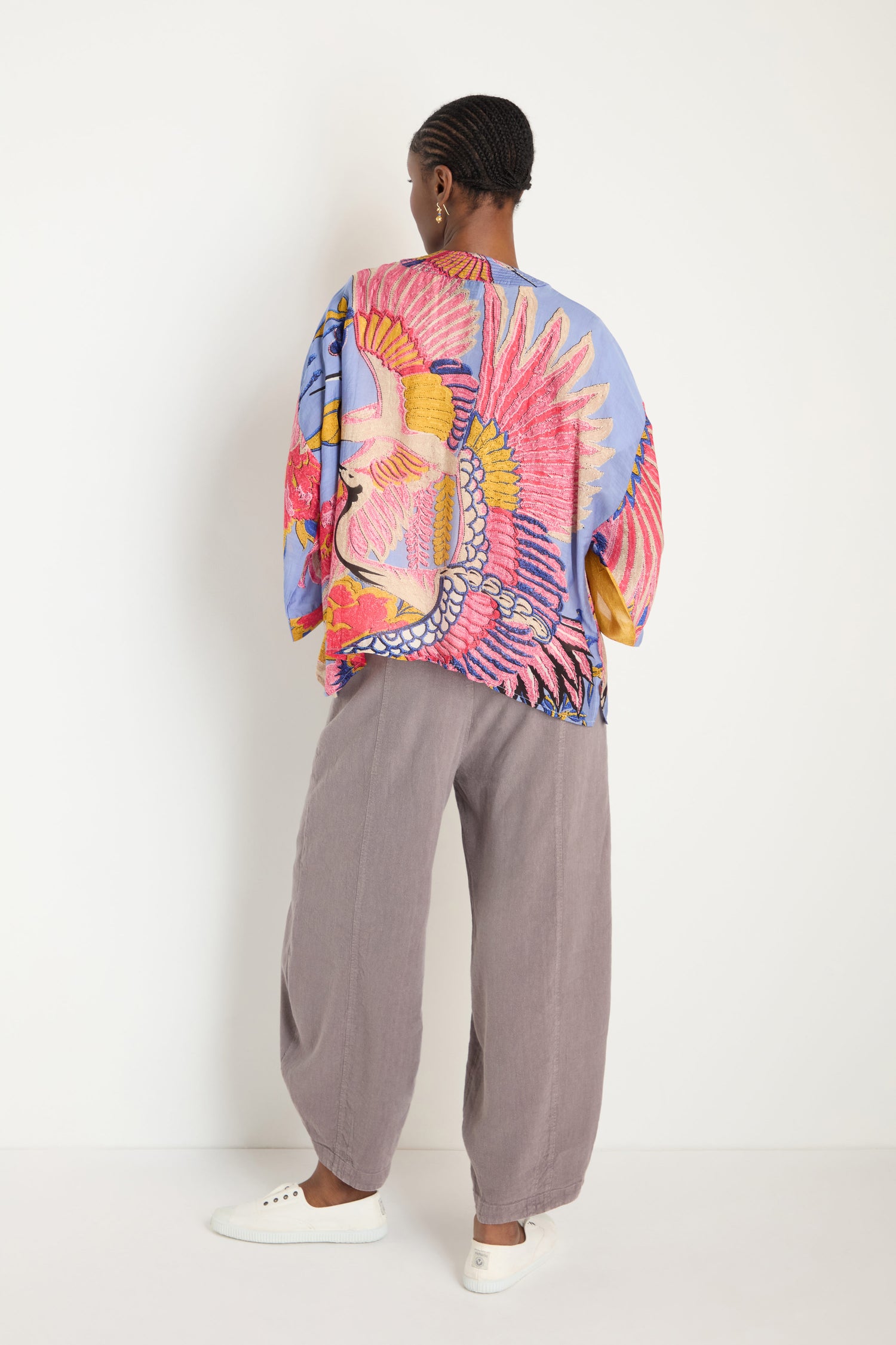 A person faces away, wearing loose grey pants, white sneakers, and a colorful oversized Crane Slate Print Kimono featuring a large bird design.