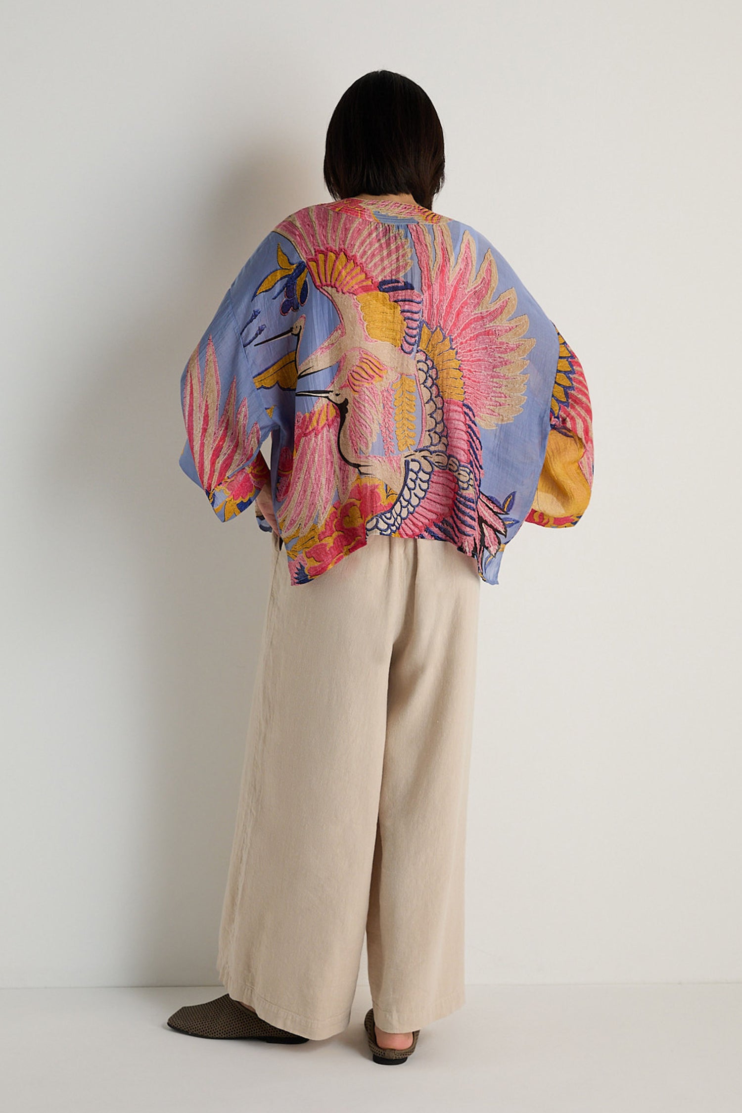 A person stands with their back to the camera, wearing a vibrant Crane Slate Print Kimono and wide beige pants, set against a plain light-colored wall.