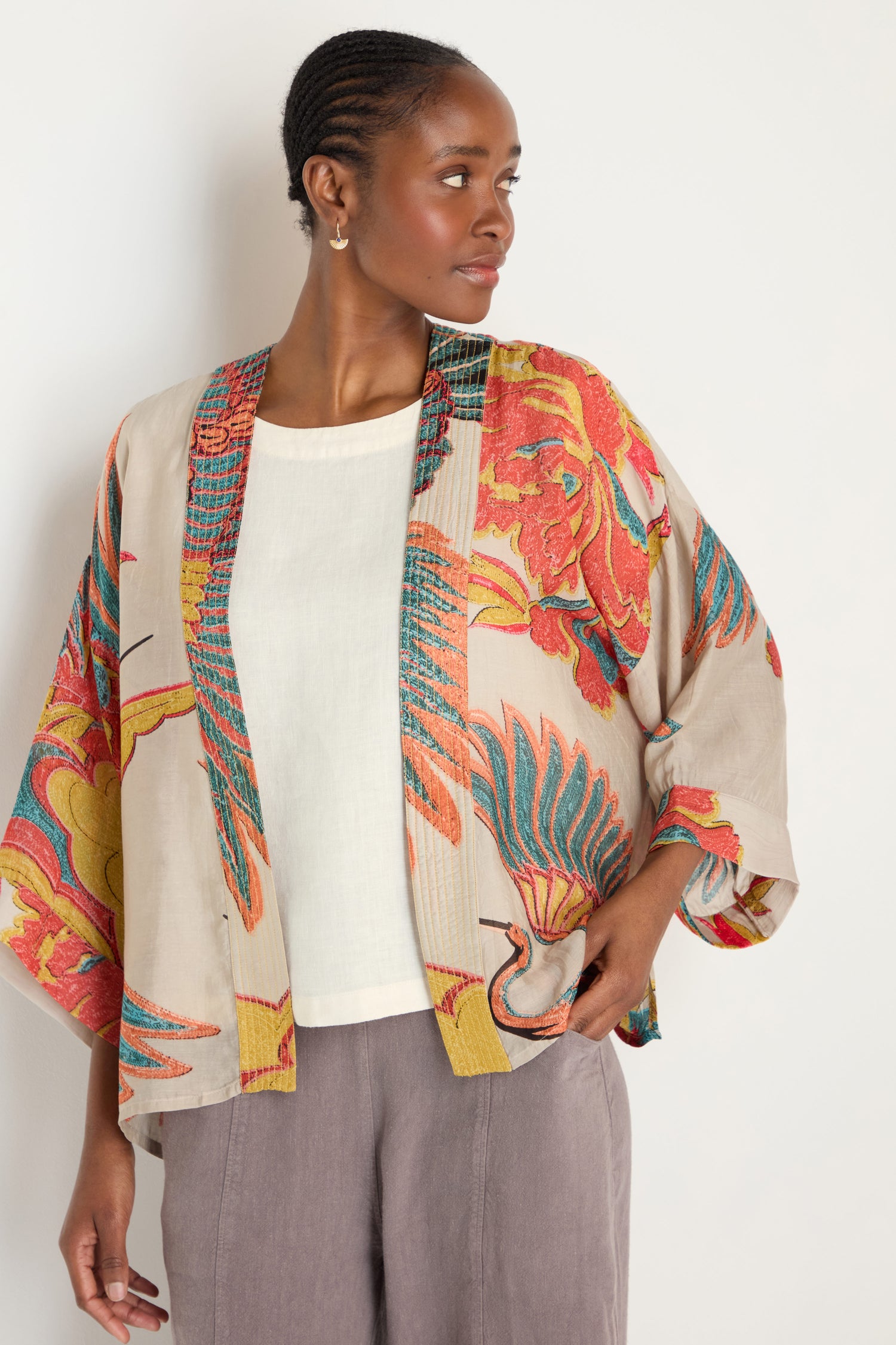 A woman in a cream top, light brown pants, and the Crane Stone Print Kimono with timeless colorful bird and floral prints stands against a plain background.