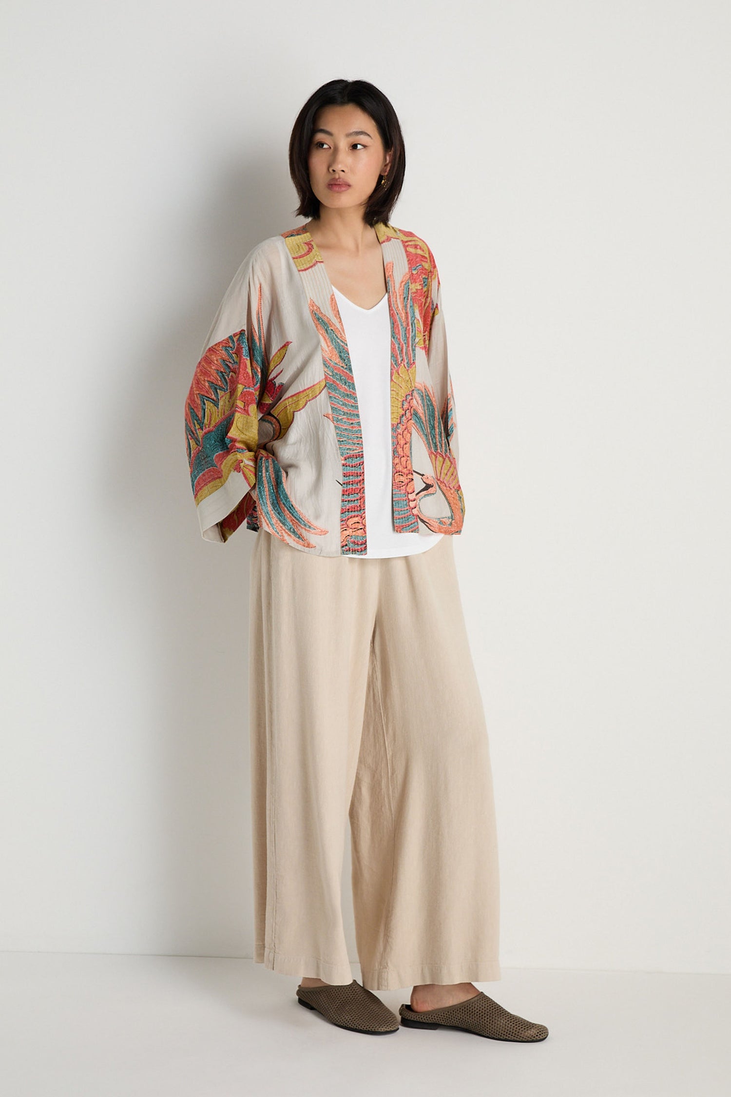 A woman stands against a white wall wearing a colorful Crane Stone Print Kimono, a white top, wide beige pants, and brown slip-on shoes.