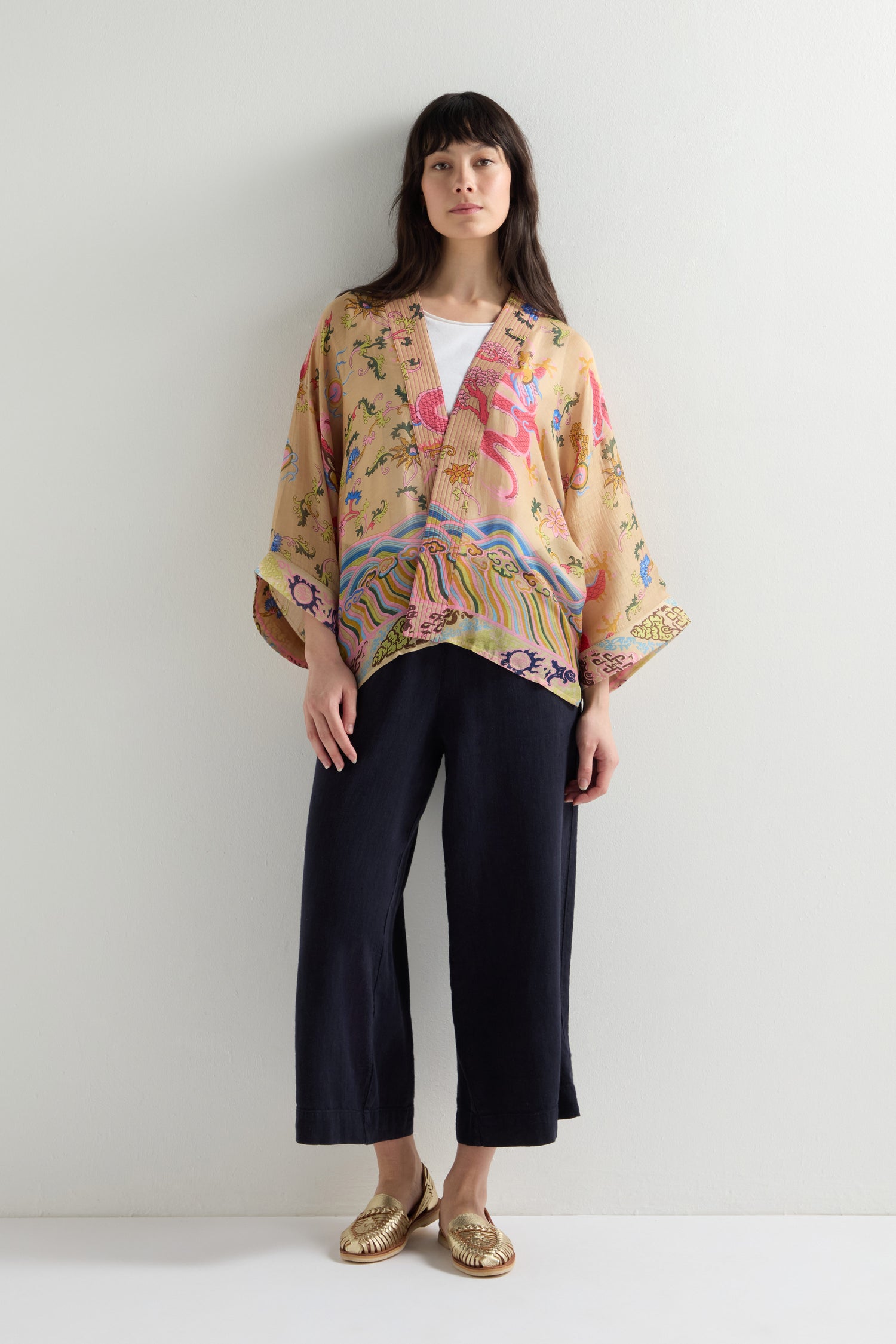 A person stands against a plain wall wearing the Dragon Print Kimono with a vintage-inspired pattern, paired with dark wide-legged pants and gold shoes.