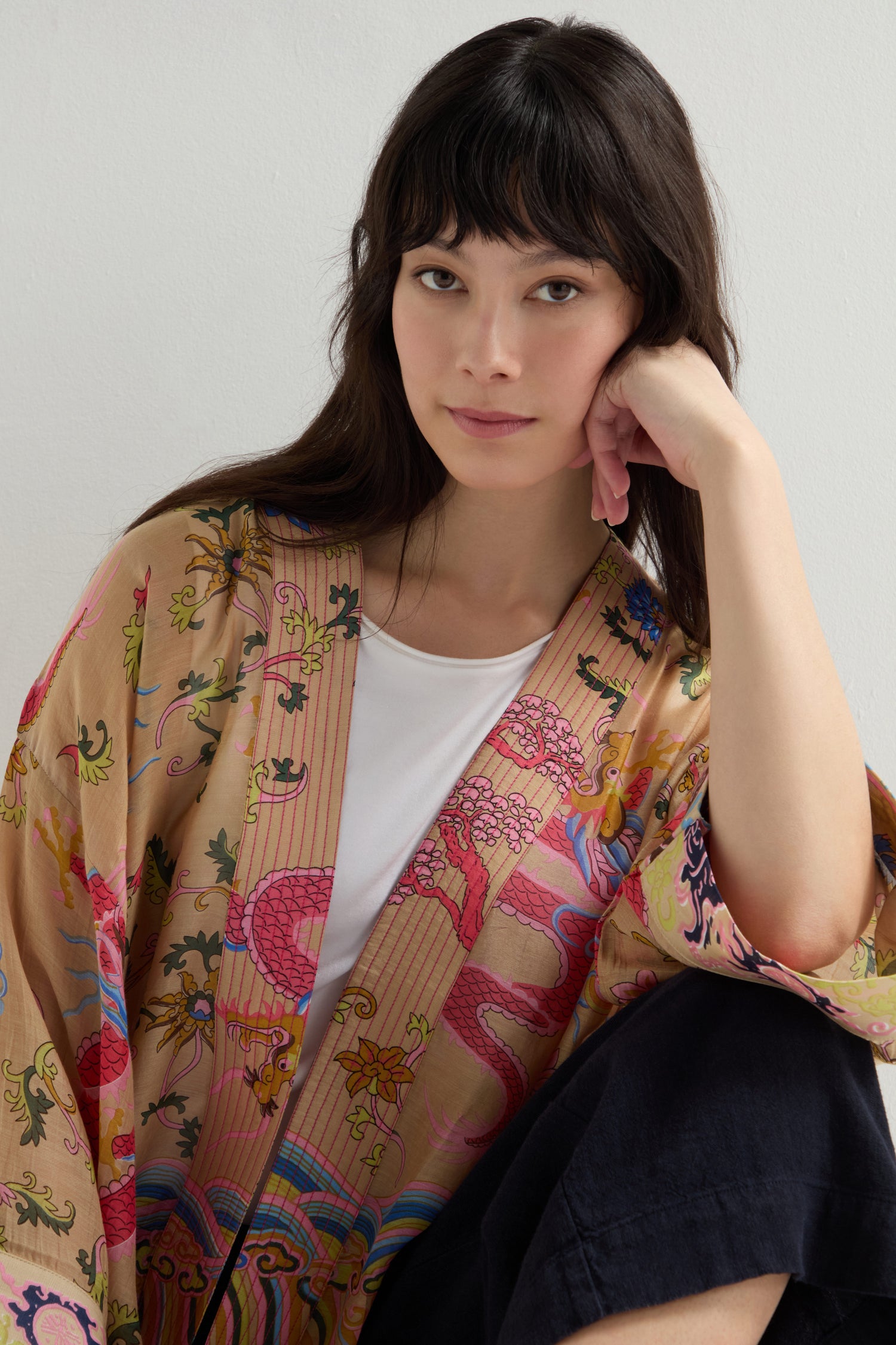 A woman with long dark hair sits elegantly, wearing the Dragon Print Kimono over a white shirt. She rests her chin on her hand, looking intently at the camera.